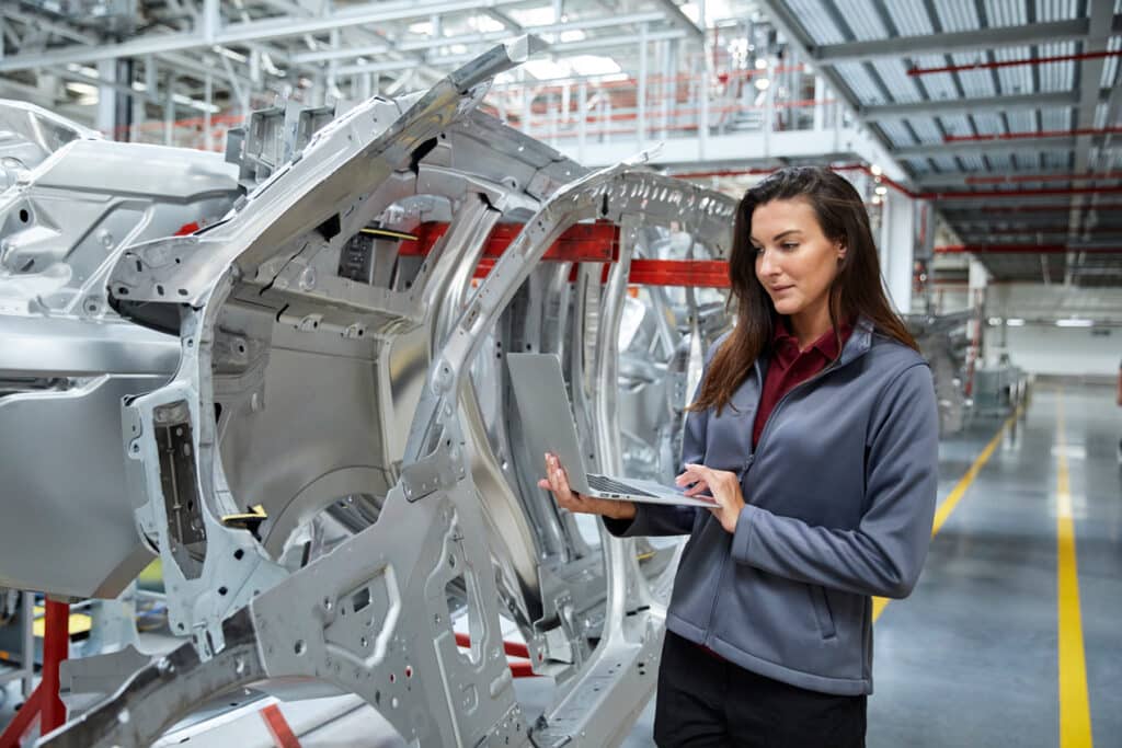 A maintenance engineer uses laptop by car chassis in automobile factory to monitor status and identify issues.