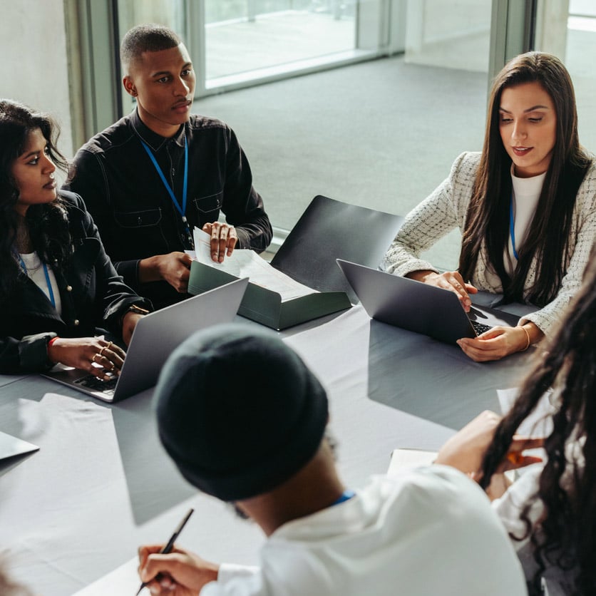 Five people discuss change management consulting at a table with laptops in a modern office.