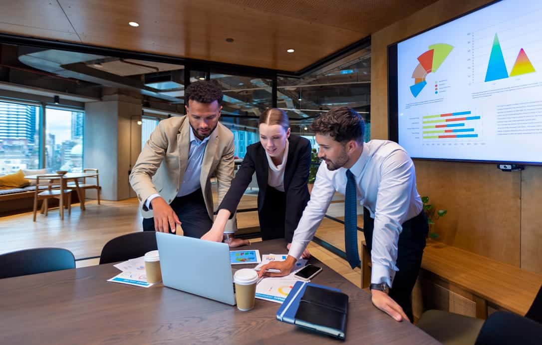 Business people working on a laptop computer in a modern office. There is paperwork on the table with charts and graphs on it. There is a screen behind them with financial charts and graphs on it.