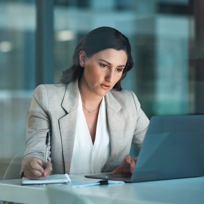 Woman in a blazer works on laptop, taking notes for Antitrust Economic Consulting at her desk.
