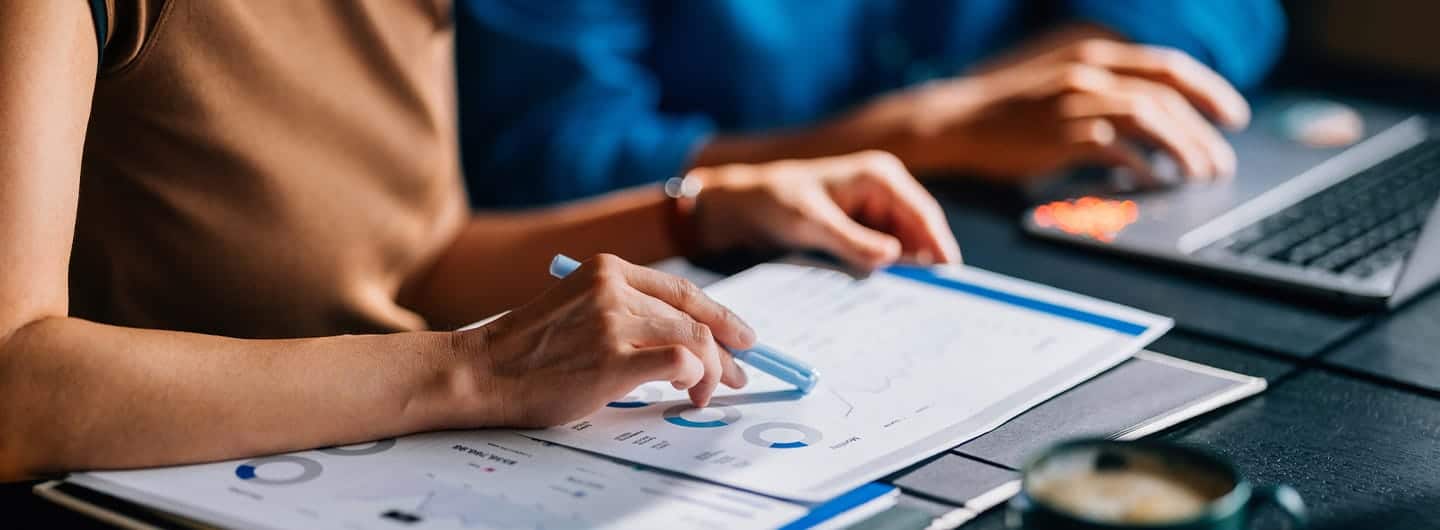 Two colleagues reviewing financial data and charts. The focus is on teamwork and analytical skills in a modern office setting with a laptop and coffee.