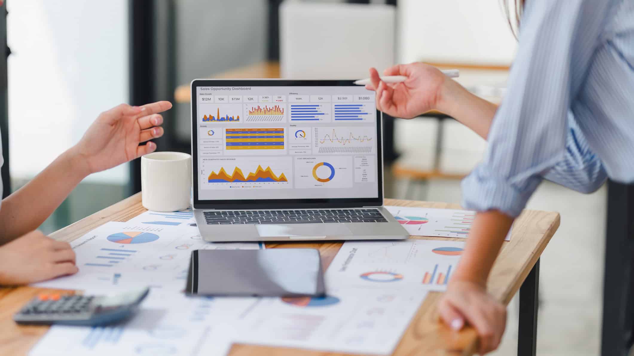 Two people discuss Customer Journey Analytics on a laptop amid documents and a tablet.