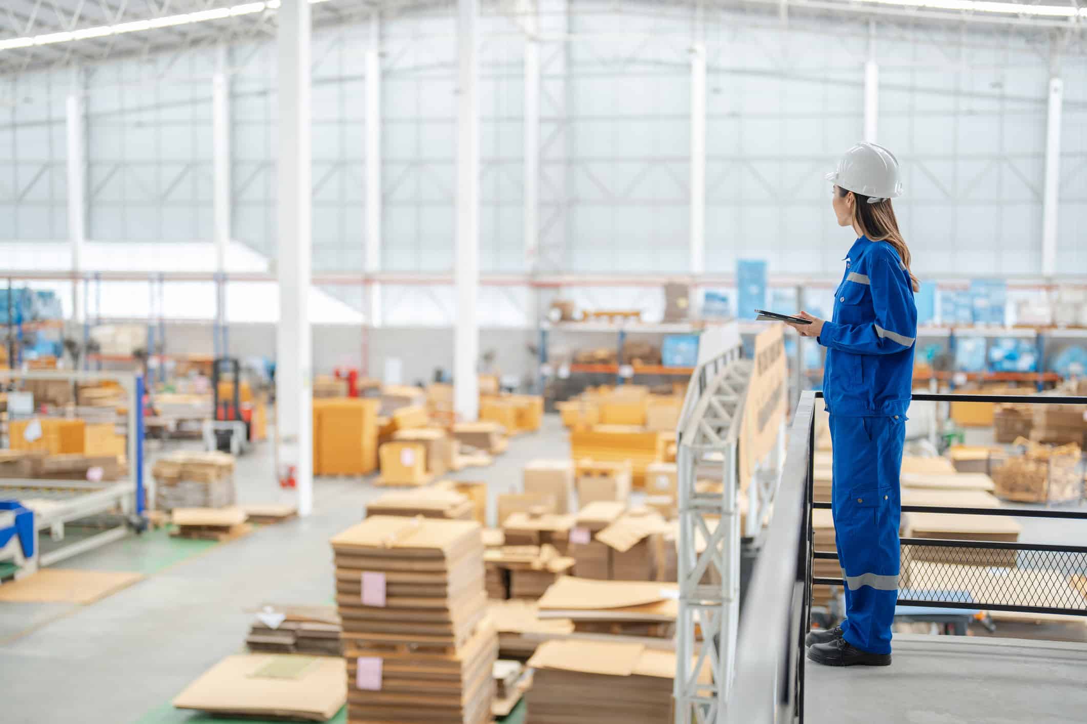 Woman in blue coveralls and helmet oversees warehouse for global logistic digital transformation.