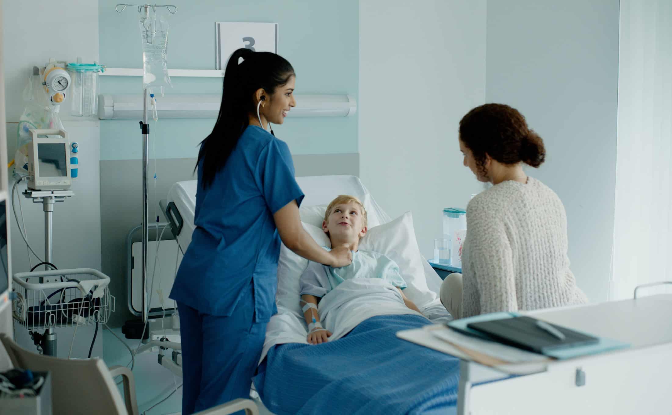 Nurse checking a young patient’s heartbeat as a woman sits by the childs hospital bed.