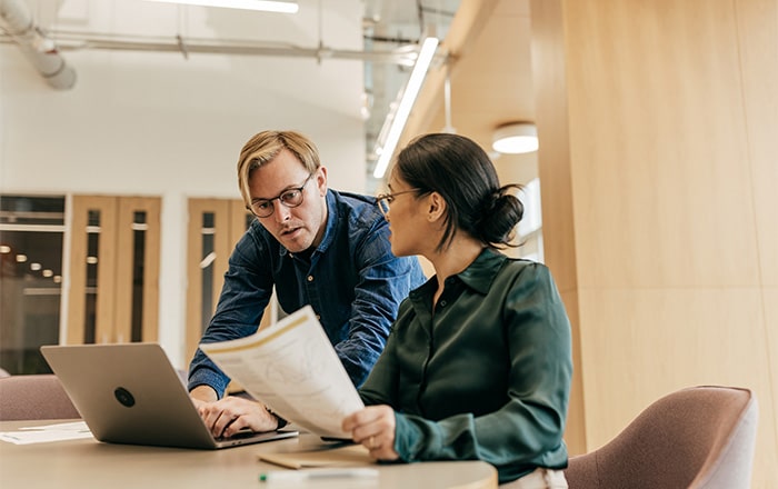 Two colleagues discuss employee benefits consulting at a desk with a laptop in a modern office.