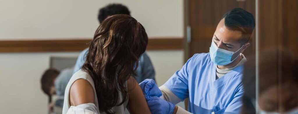 A healthcare worker in a mask gives a vaccination to a seated patient in a clinic.