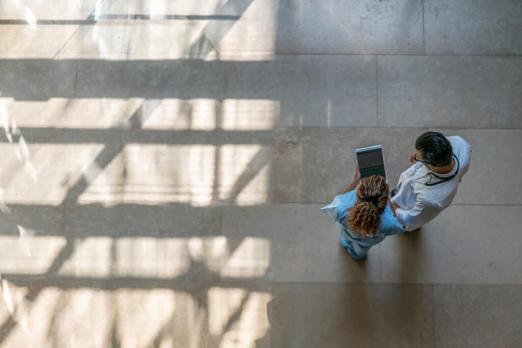 Two medical professionals view a tablet while standing in a sunlit hallway, seen from above.