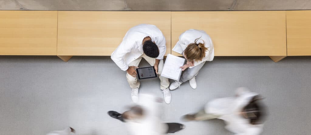 Two people in white coats sit on a bench, looking at a tablet and papers; others walk by.