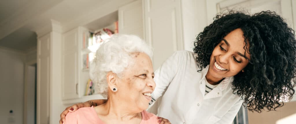 Smiling caregiver supports and chats with a happy older woman in a bright kitchen.