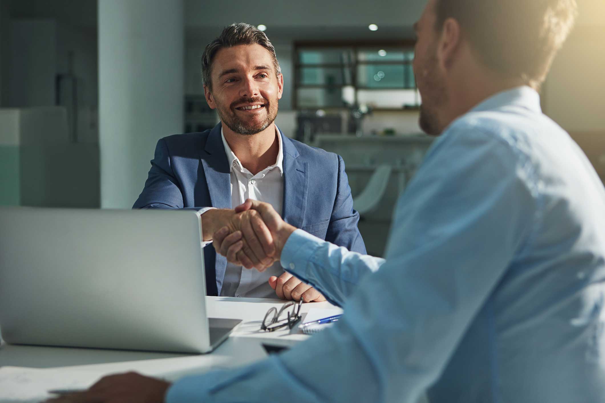 Two men in business attire shake hands across a desk, discussing certification & audit support.