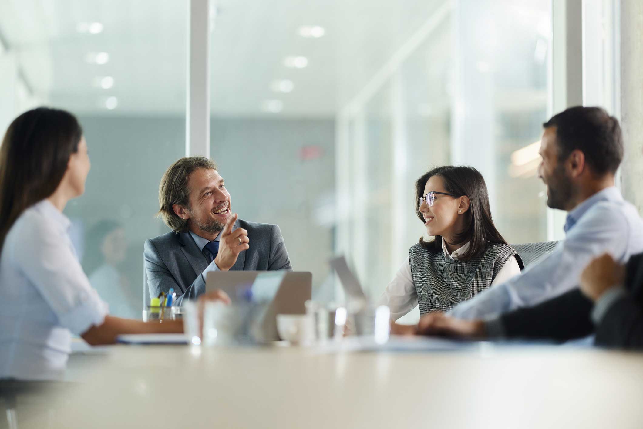 Four colleagues discuss client relationship analytics while smiling during a conference meeting.