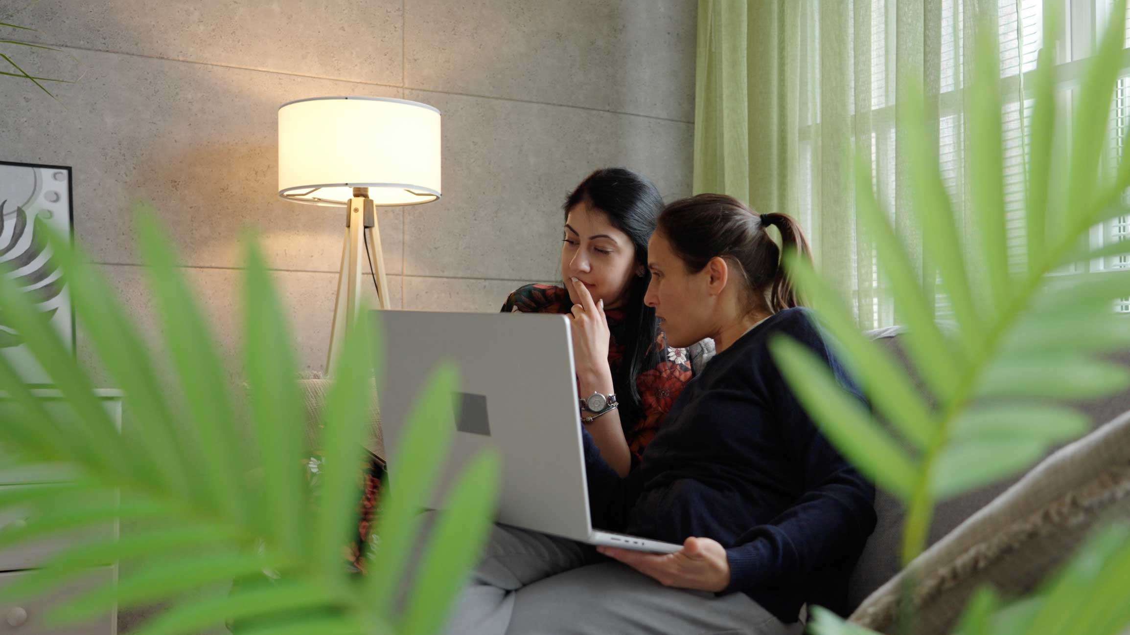 Two women discuss identity access management advisory while viewing a laptop, amid green plants.