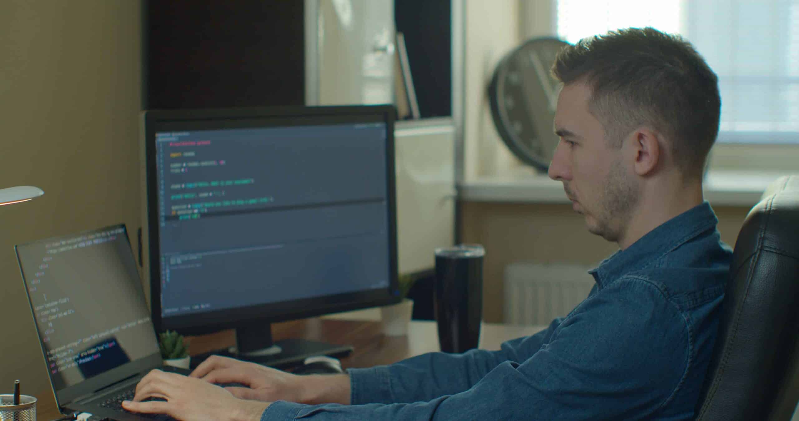 Man working at a desk with two monitors displaying code for compliance monitoring in an office.