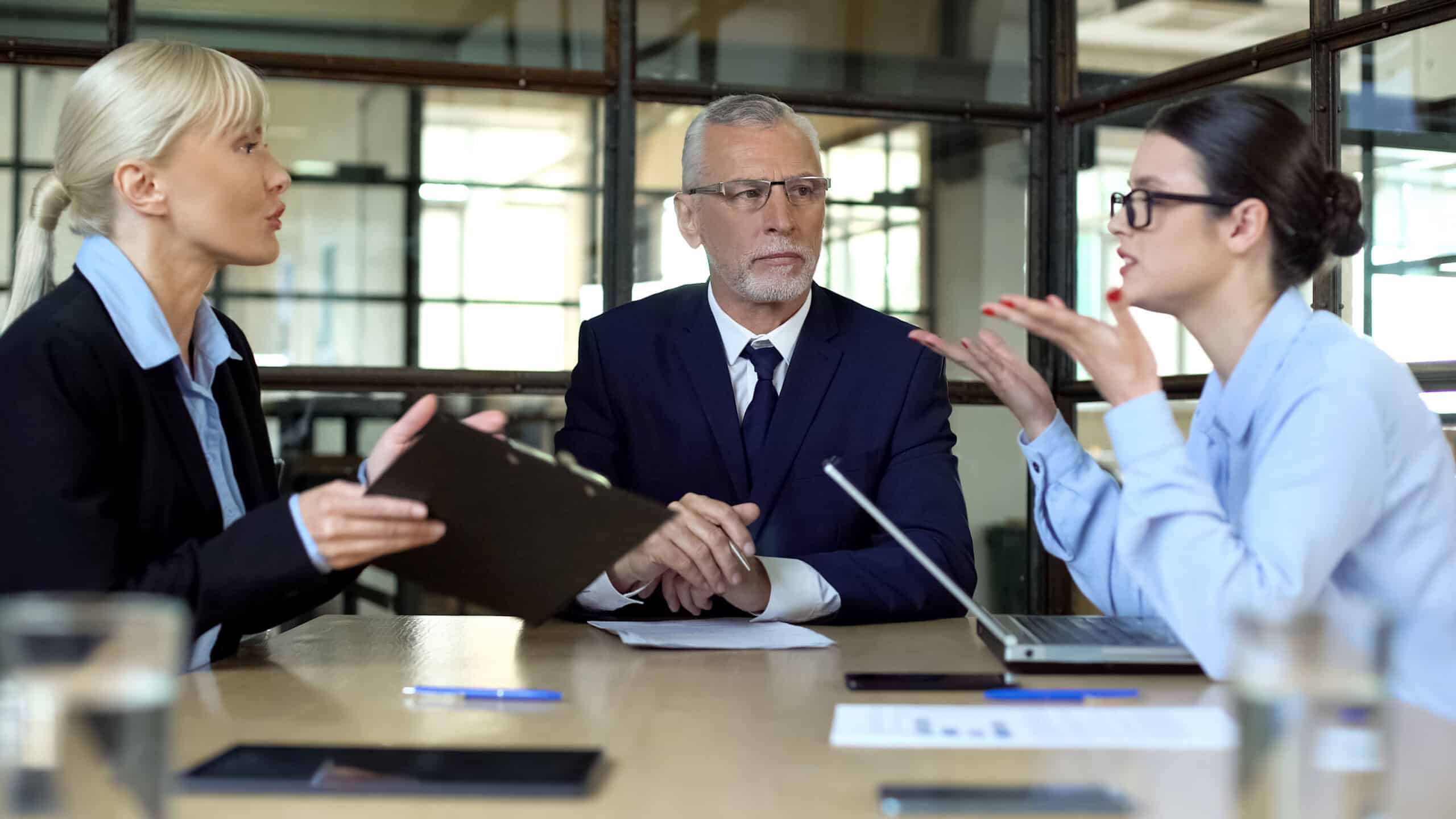 Two women argue at table while a man, seeking Big Data Strategy Consulting Services, looks concerned