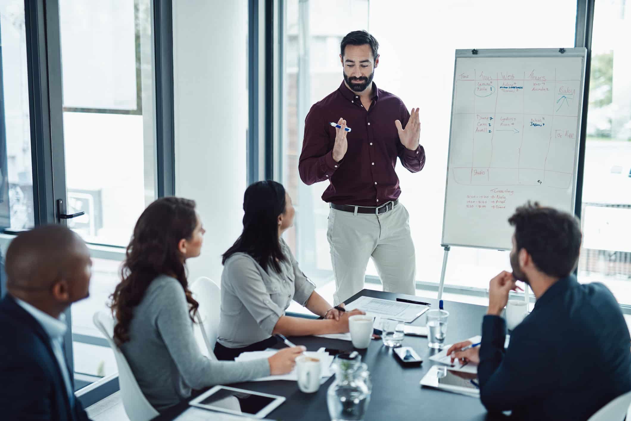 A man presents Business Intelligence Optimization at a whiteboard to colleagues in a modern office.