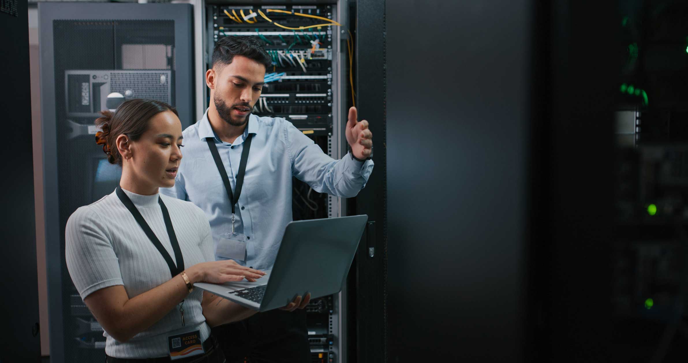 Two email security consultants work with a laptop in a server room full of network equipment.