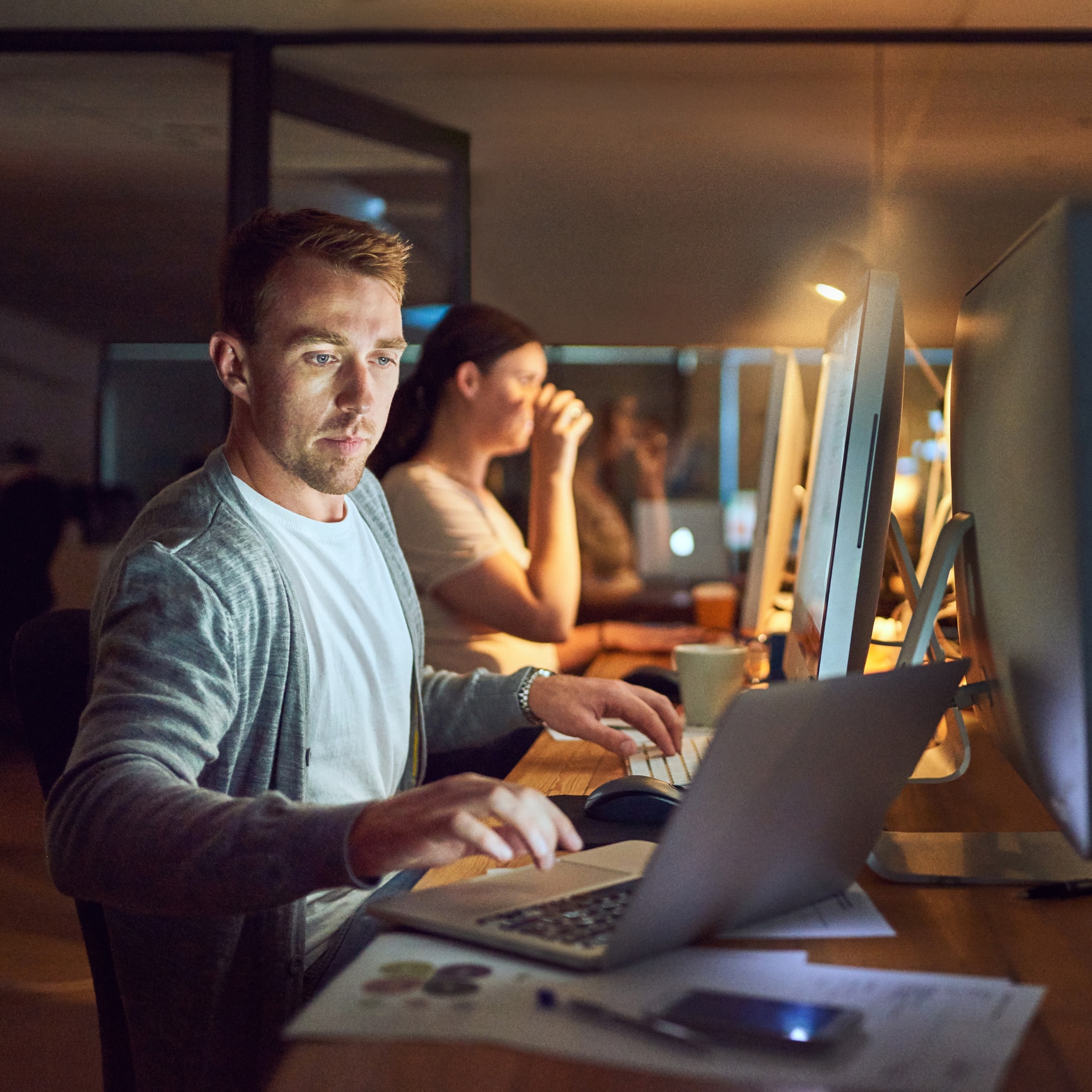 Man working at computer in dark office, enabling competitive strategy; woman drinks from a mug.
