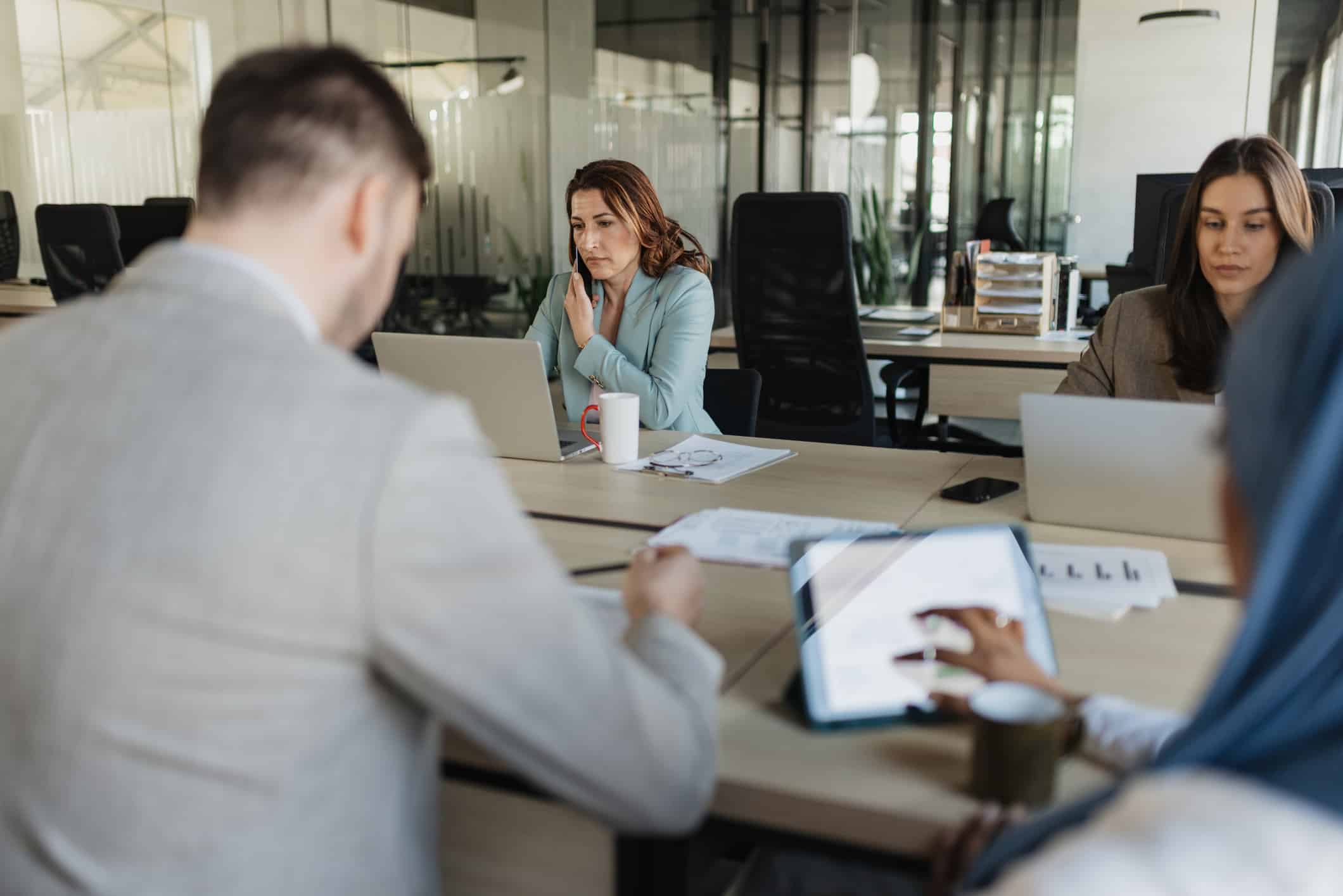 Four people discuss Competitive Strategy Advisory at a shared office table with laptops and coffee.