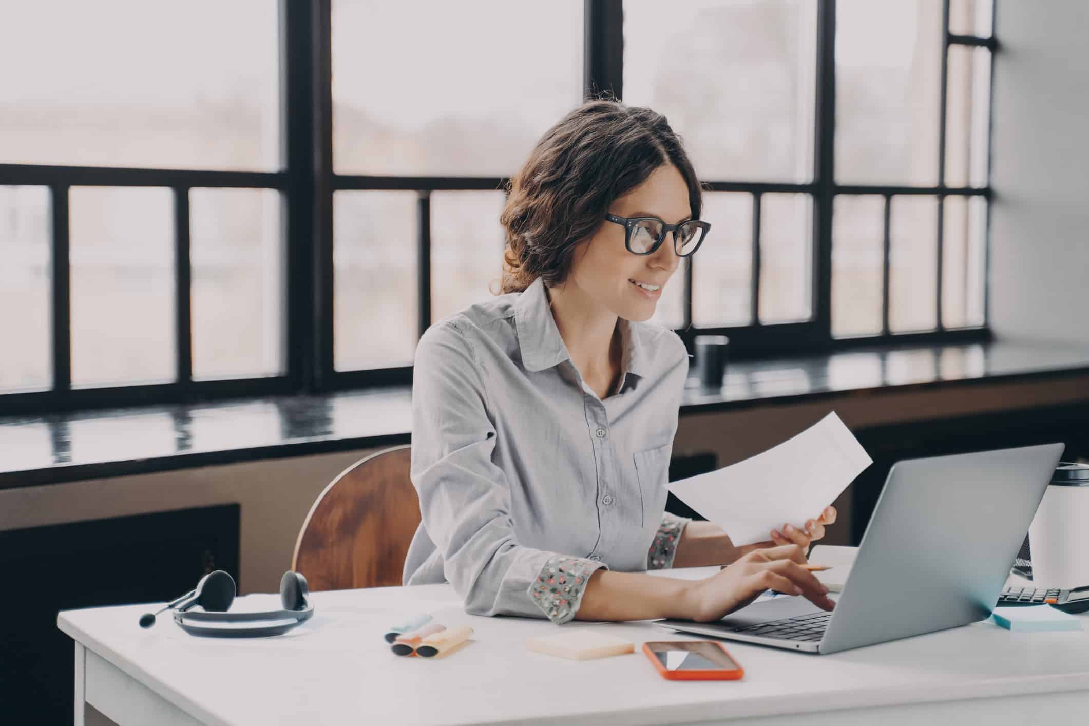 Woman with glasses prepares for witness preparation on laptop, holding a paper and smiling.