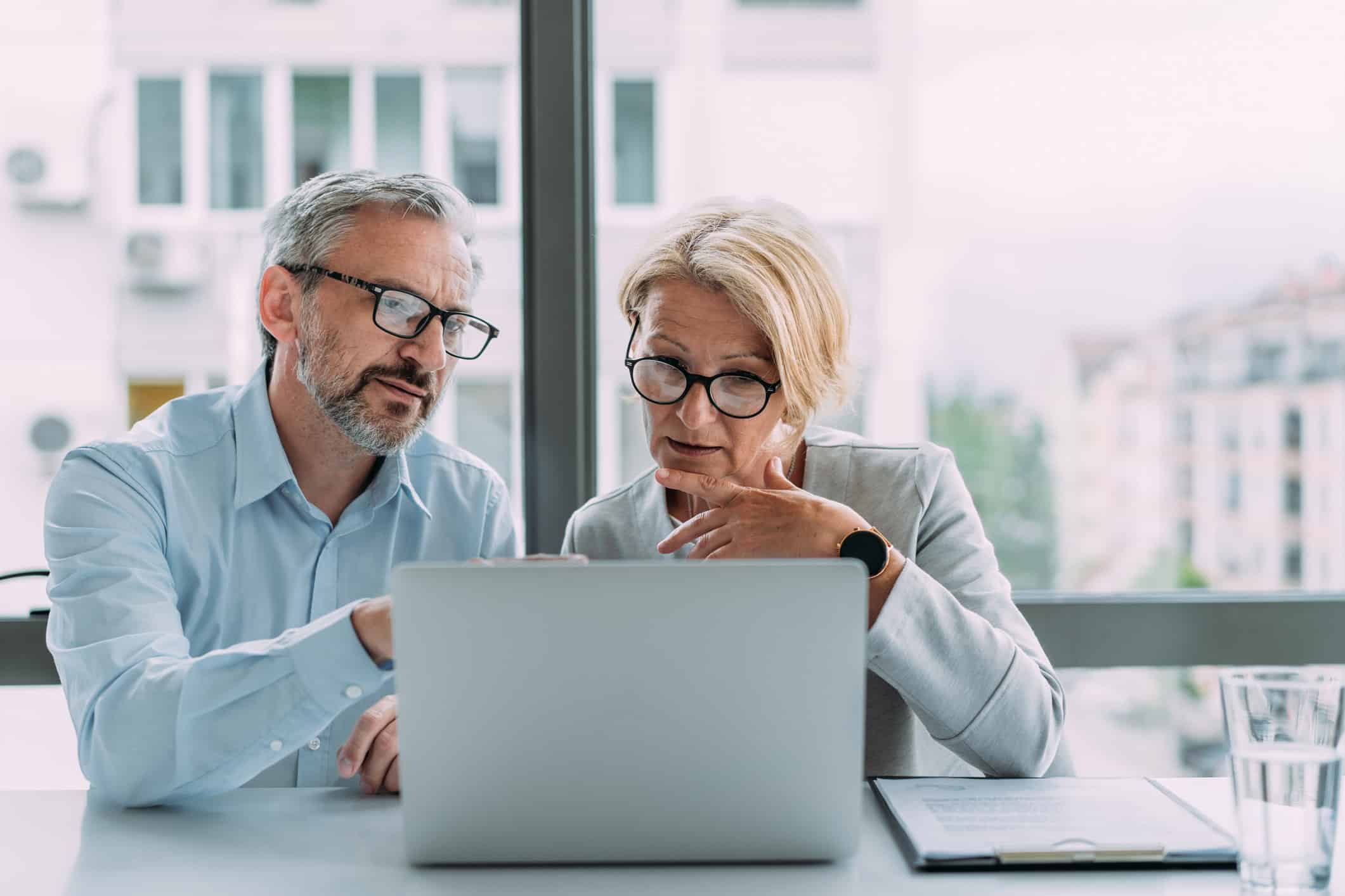 Two older adults with glasses review documents at a law firm in a bright office together.