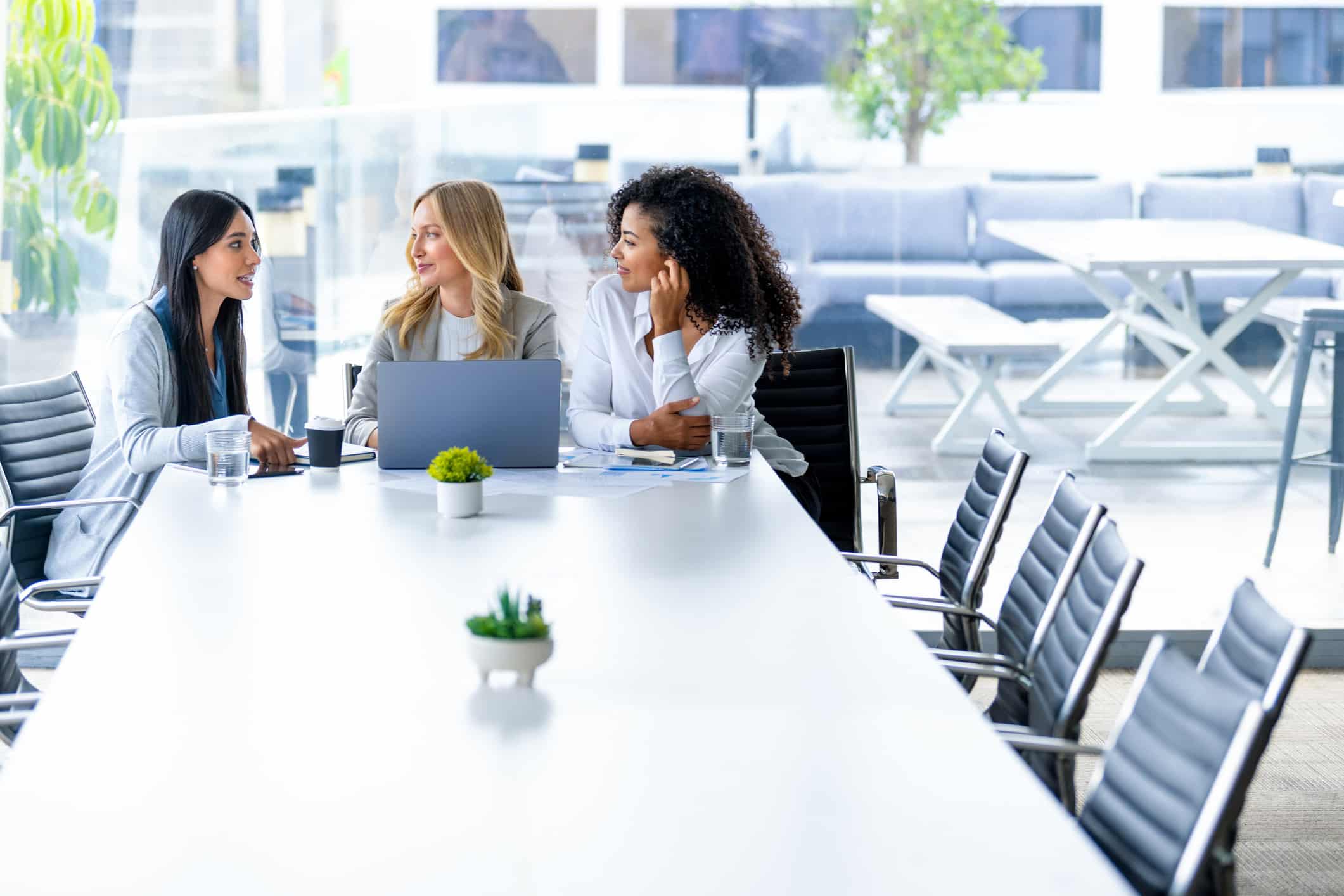 Three women discuss documents and a laptop at a Law Firm Growth Consulting Services meeting.
