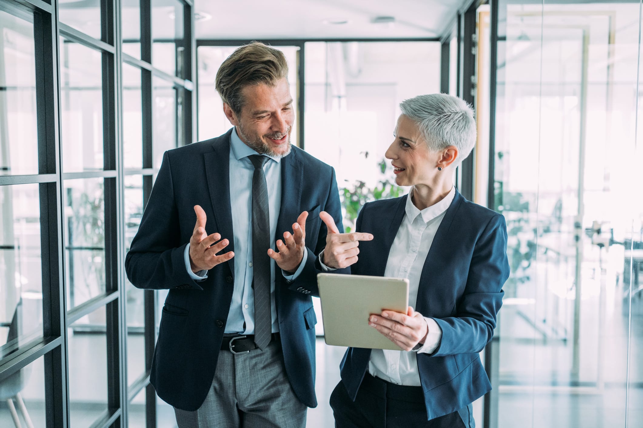 Two businesspeople discuss Law Firm Growth Execution while smiling at a tablet in office hallway.
