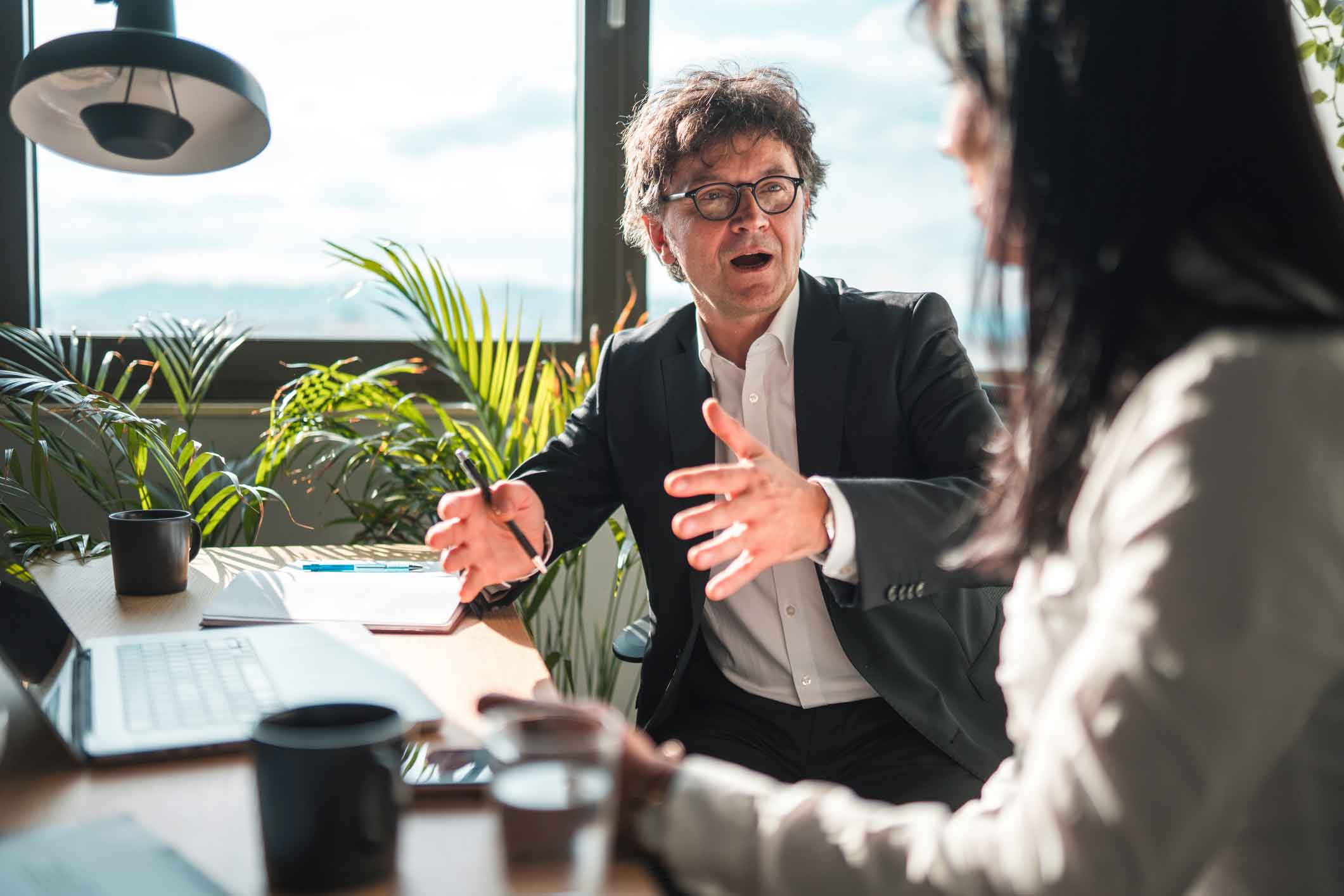 Man in suit discussing cybersecurity compliance Execution with woman in bright, plant-filled office.