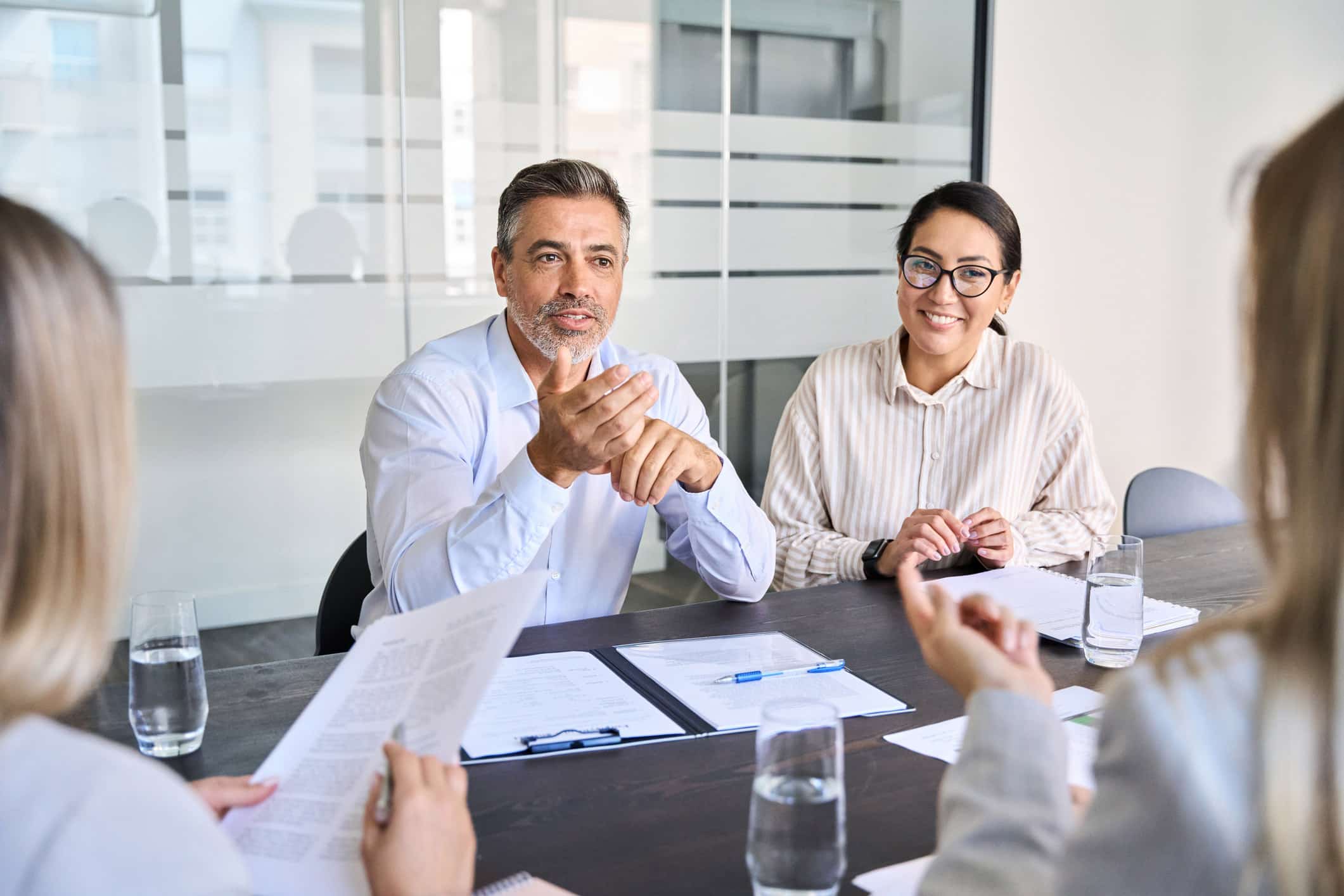 Two people at a meeting table discussing Mergers & Acquisitions Execution with documents and water.