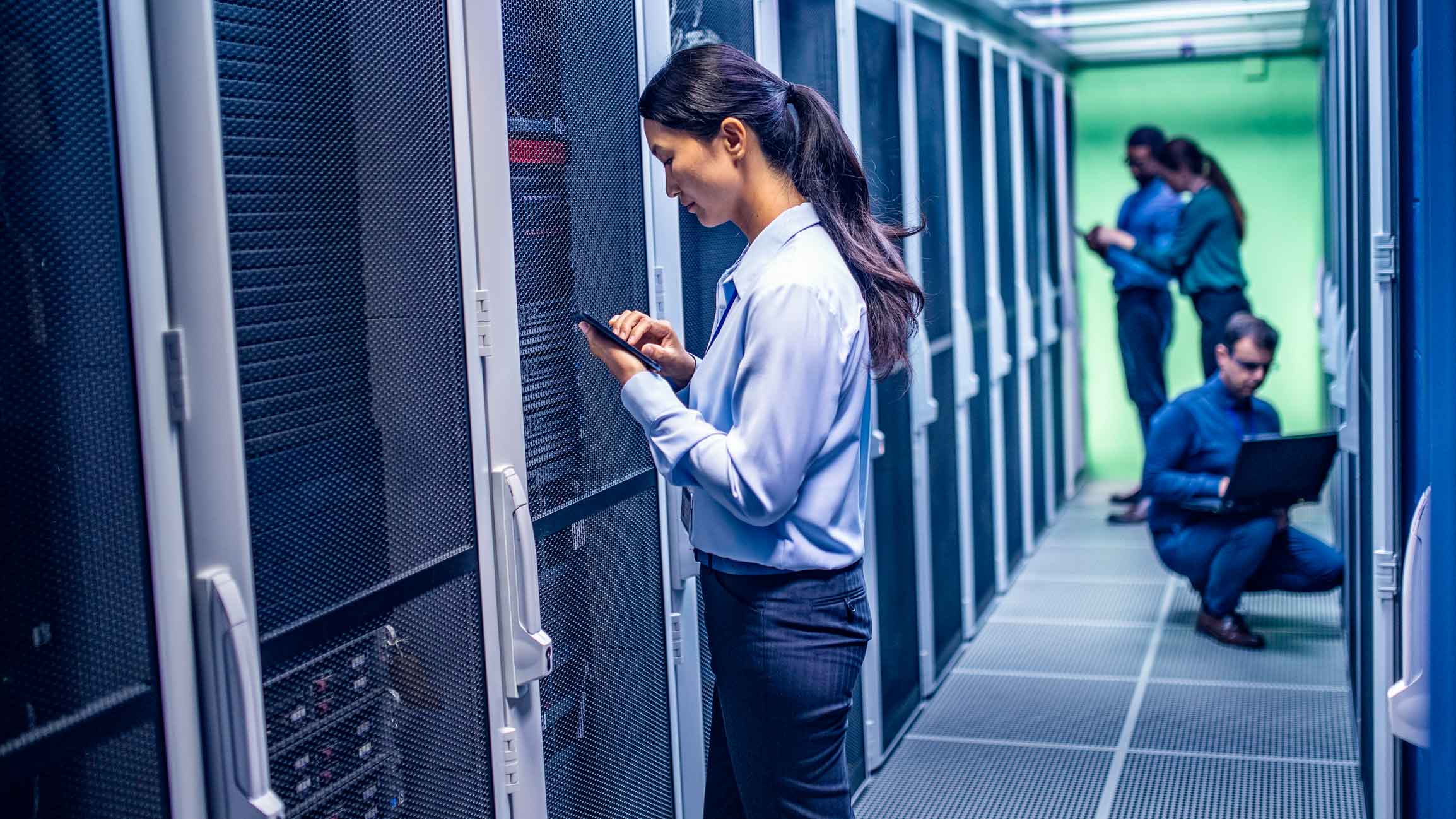 Woman using tablet in server room during a forensic investigation, with colleagues in background.