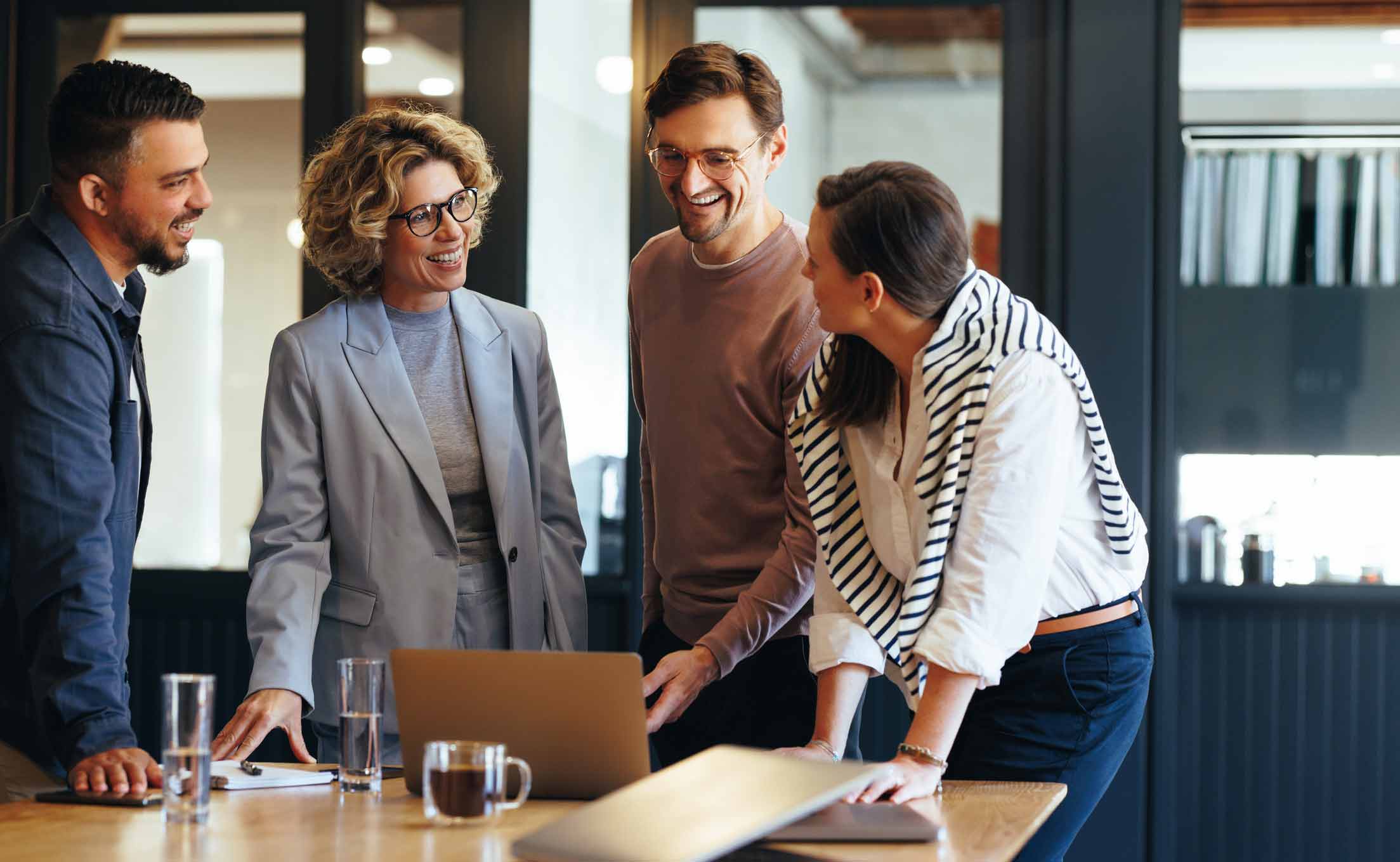Four people smiling and talking around a laptop in a modern office setting.
