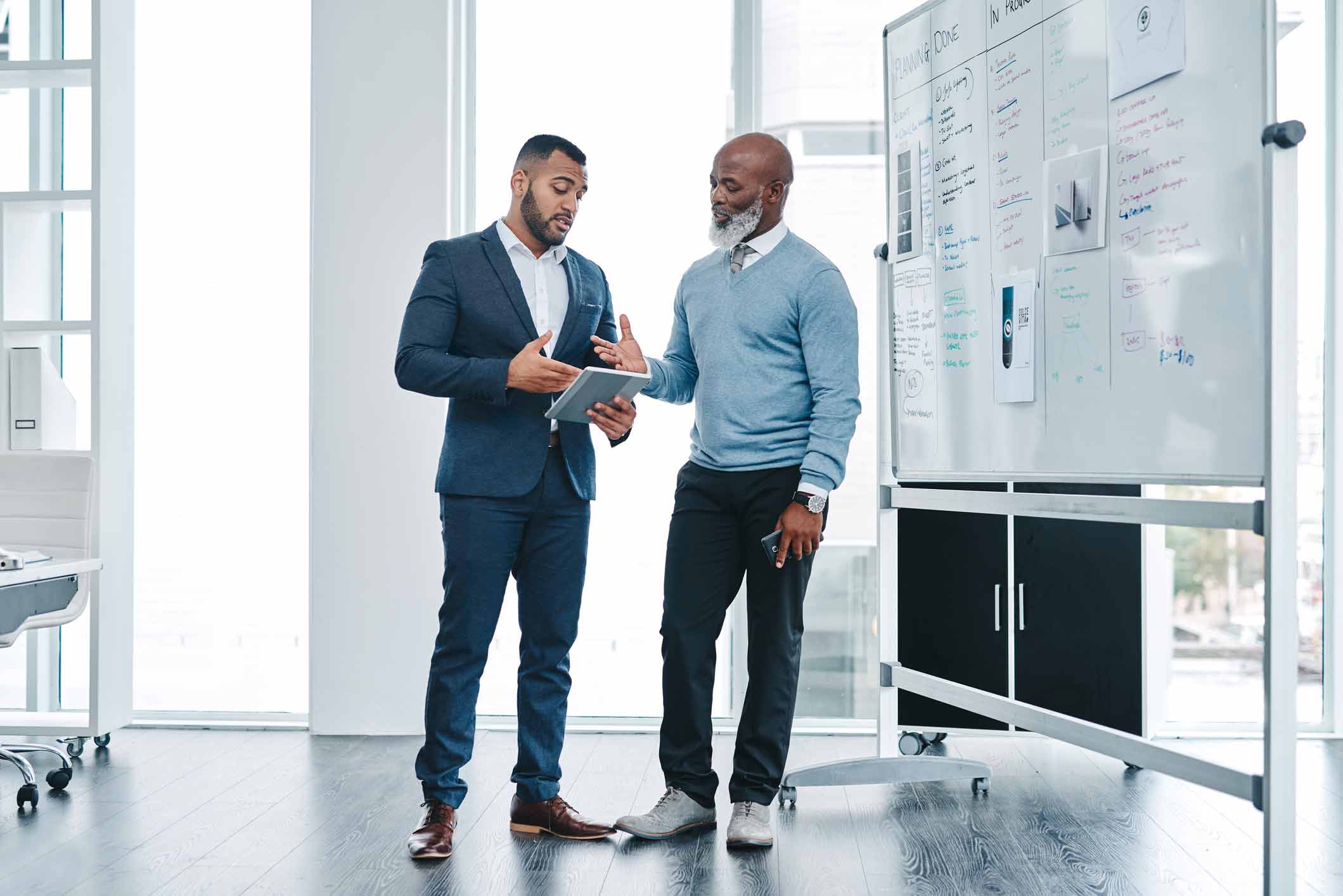 Two men discussing near a whiteboard with notes and diagrams in a modern office.