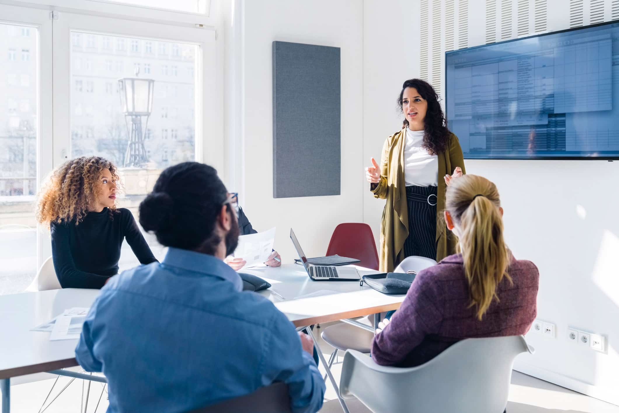 Woman presenting to colleagues in a bright office meeting room with a screen on the wall.