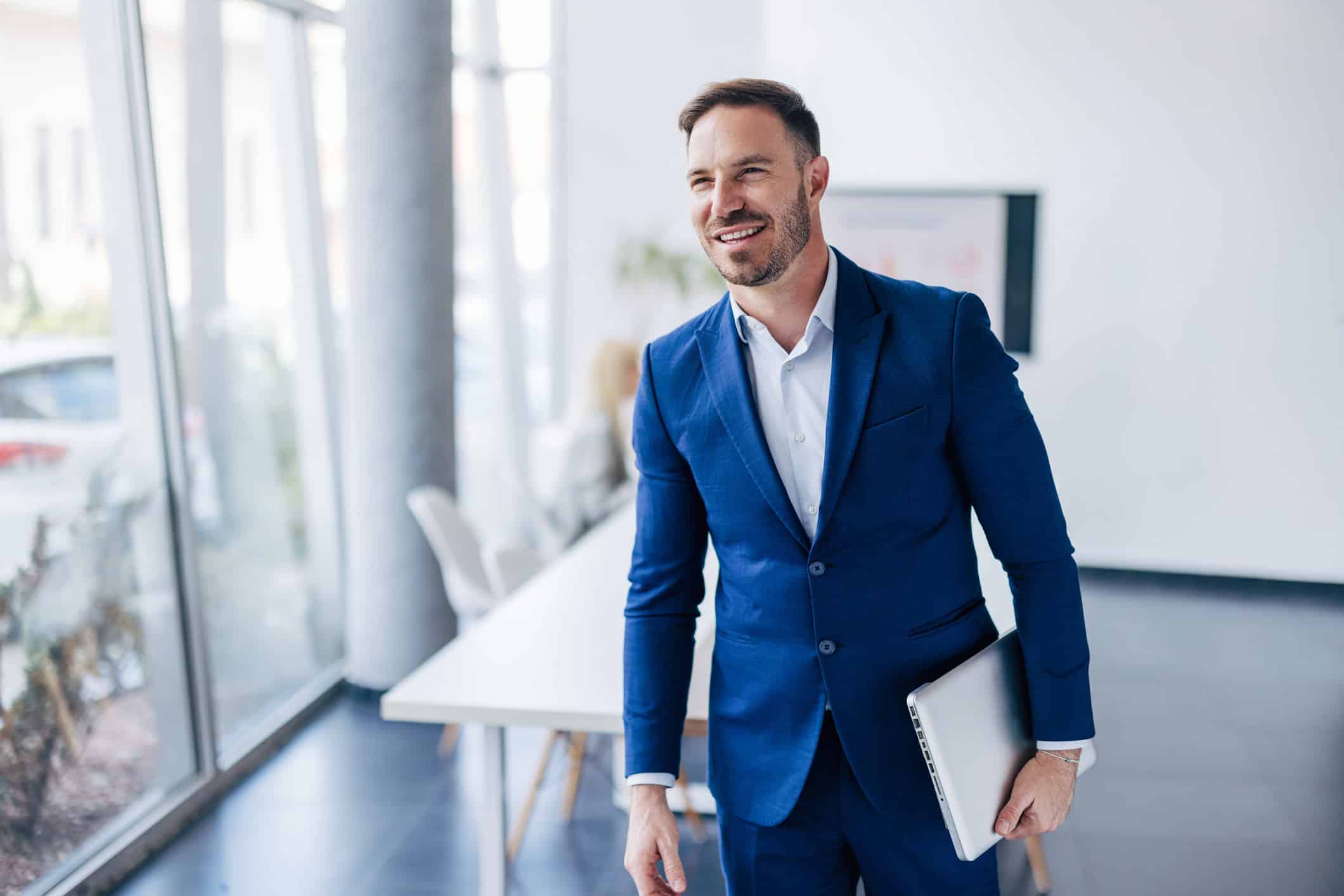 Man in blue suit smiling, holding a laptop, showcasing law firm growth strategy by the window.