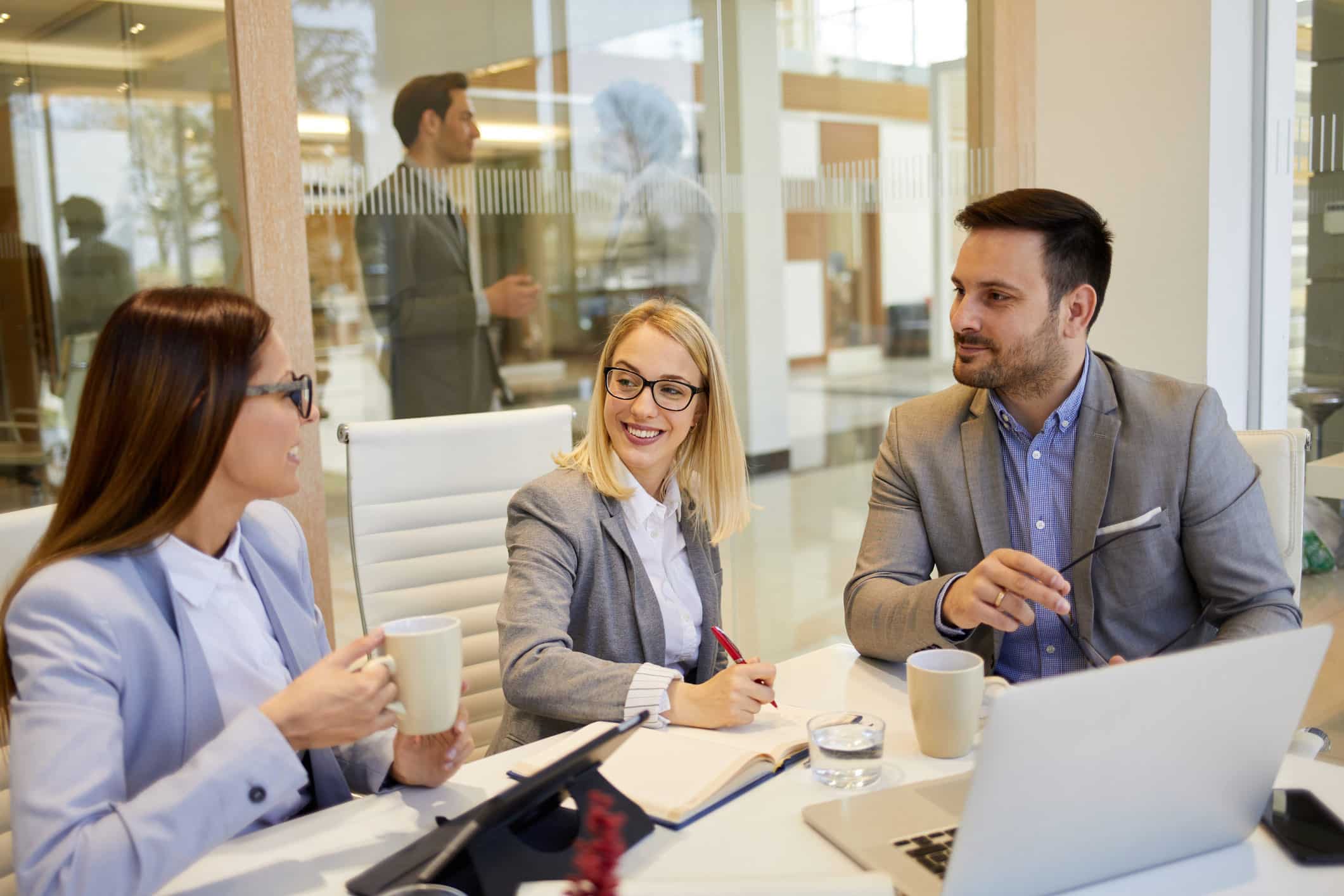 Three businesspeople discuss witness preparation at a meeting table with laptops and coffee.