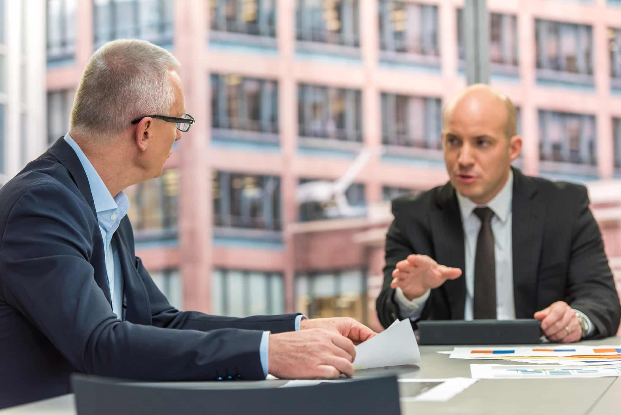 Two businessmen in suits discuss M&A Advisory documents at a table in a modern office.