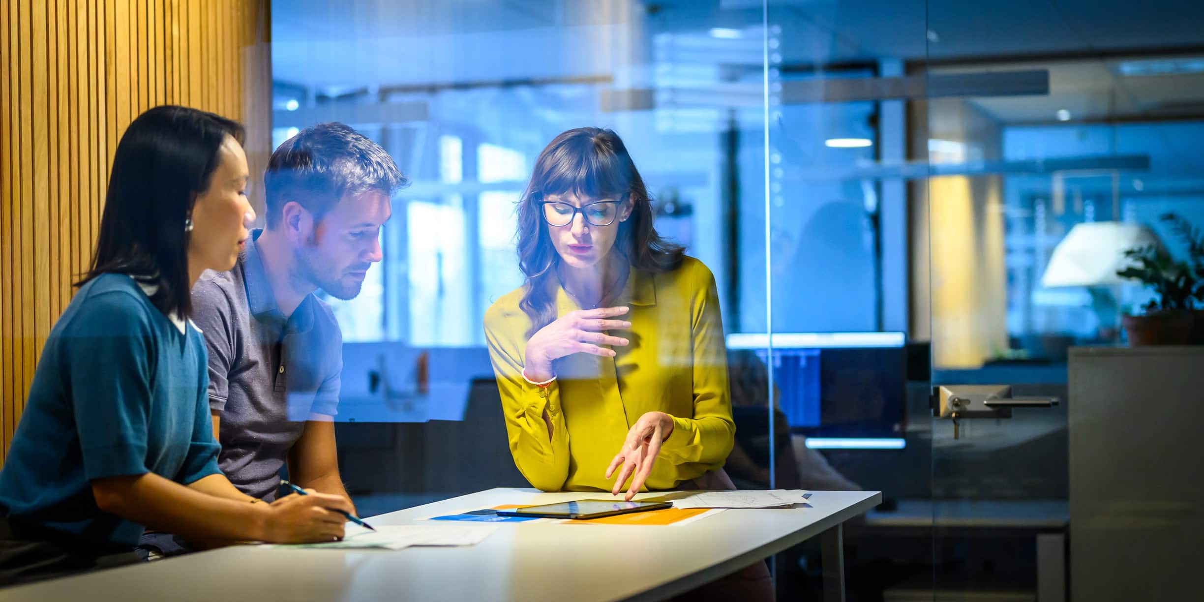 Three people sit at a table in an office, reviewing Mergers & Acquisitions Advisory documents.