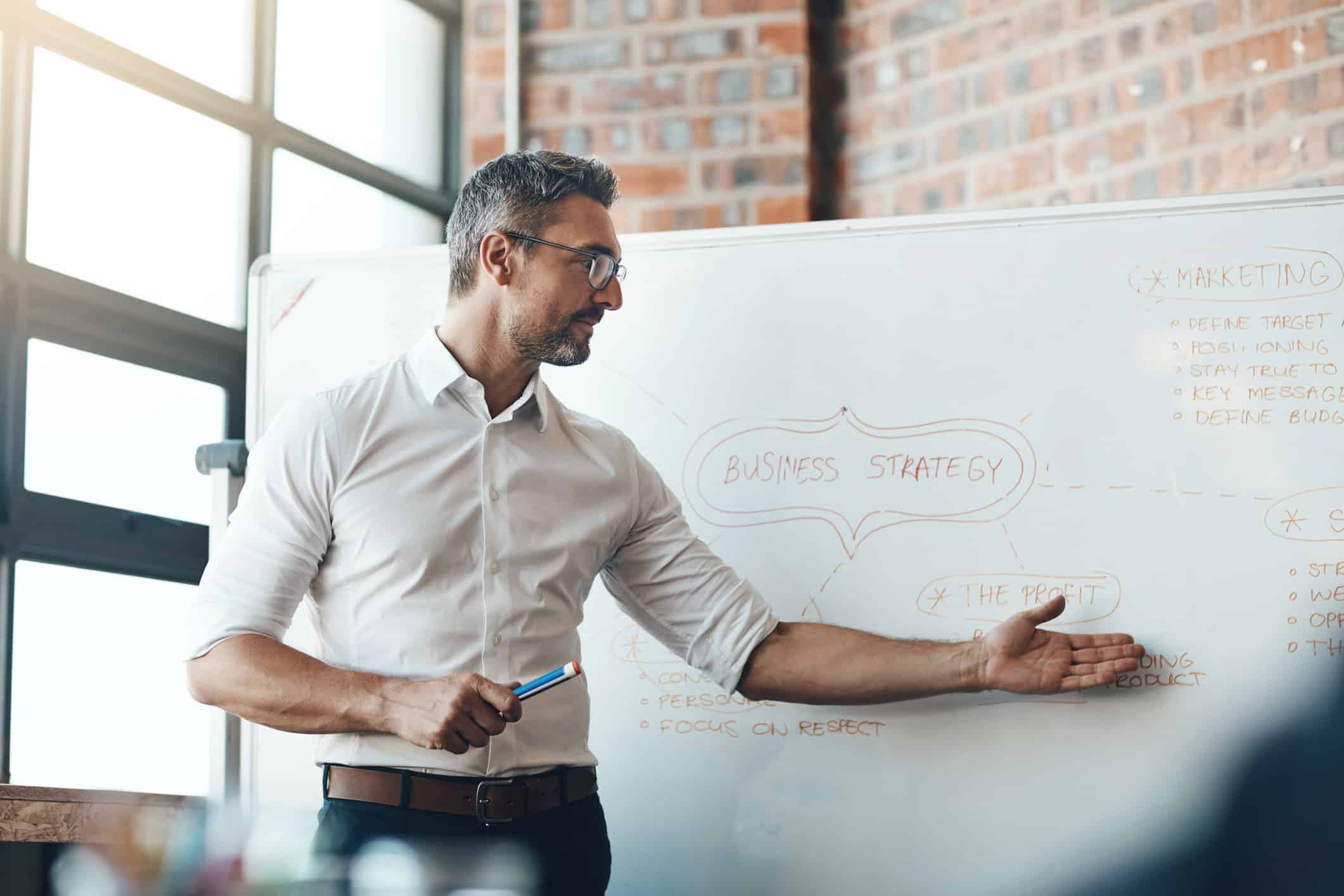 Man in glasses presenting business valuation strategy on a whiteboard in an office setting.
