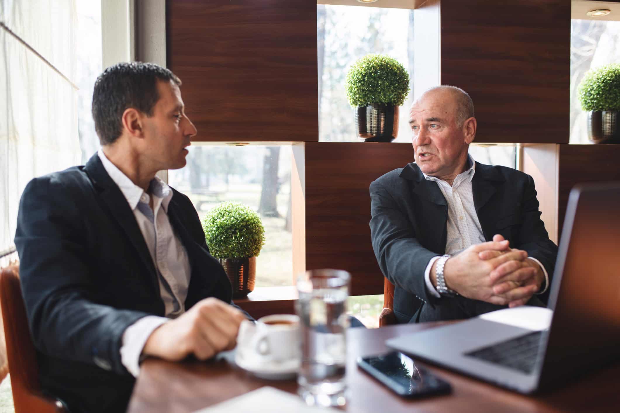 Two Risk Management Consultants in suits discuss strategy over coffee, water, and a laptop.