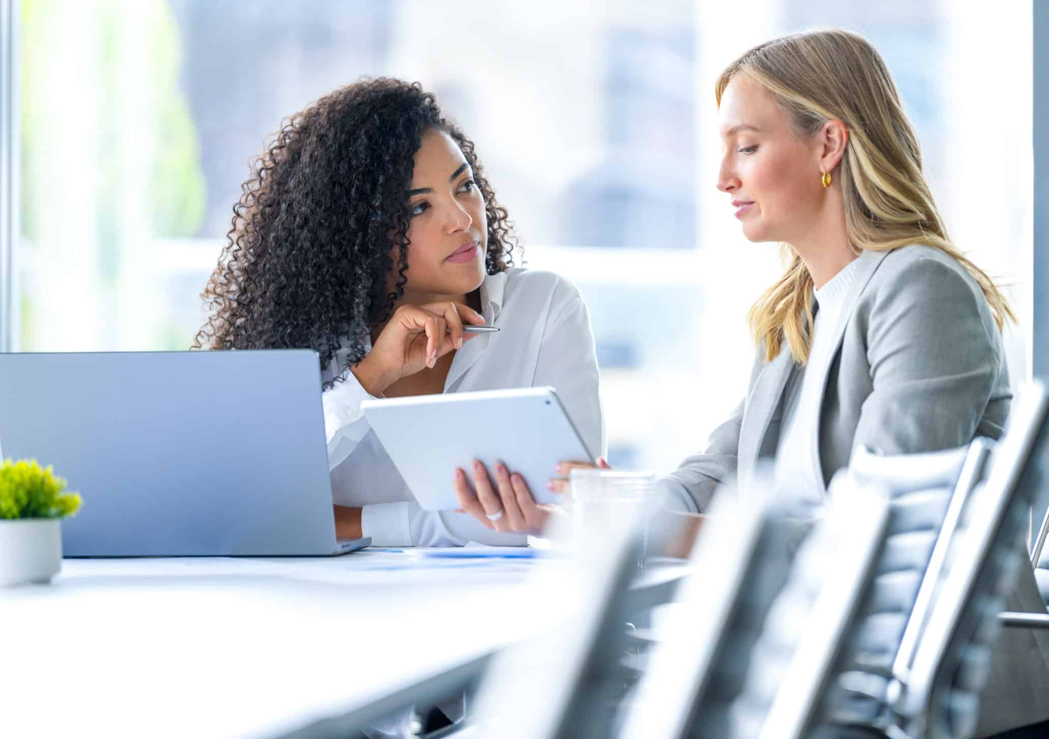 Two women in business attire discuss M&A strategy consulting firms while viewing a tablet in office.