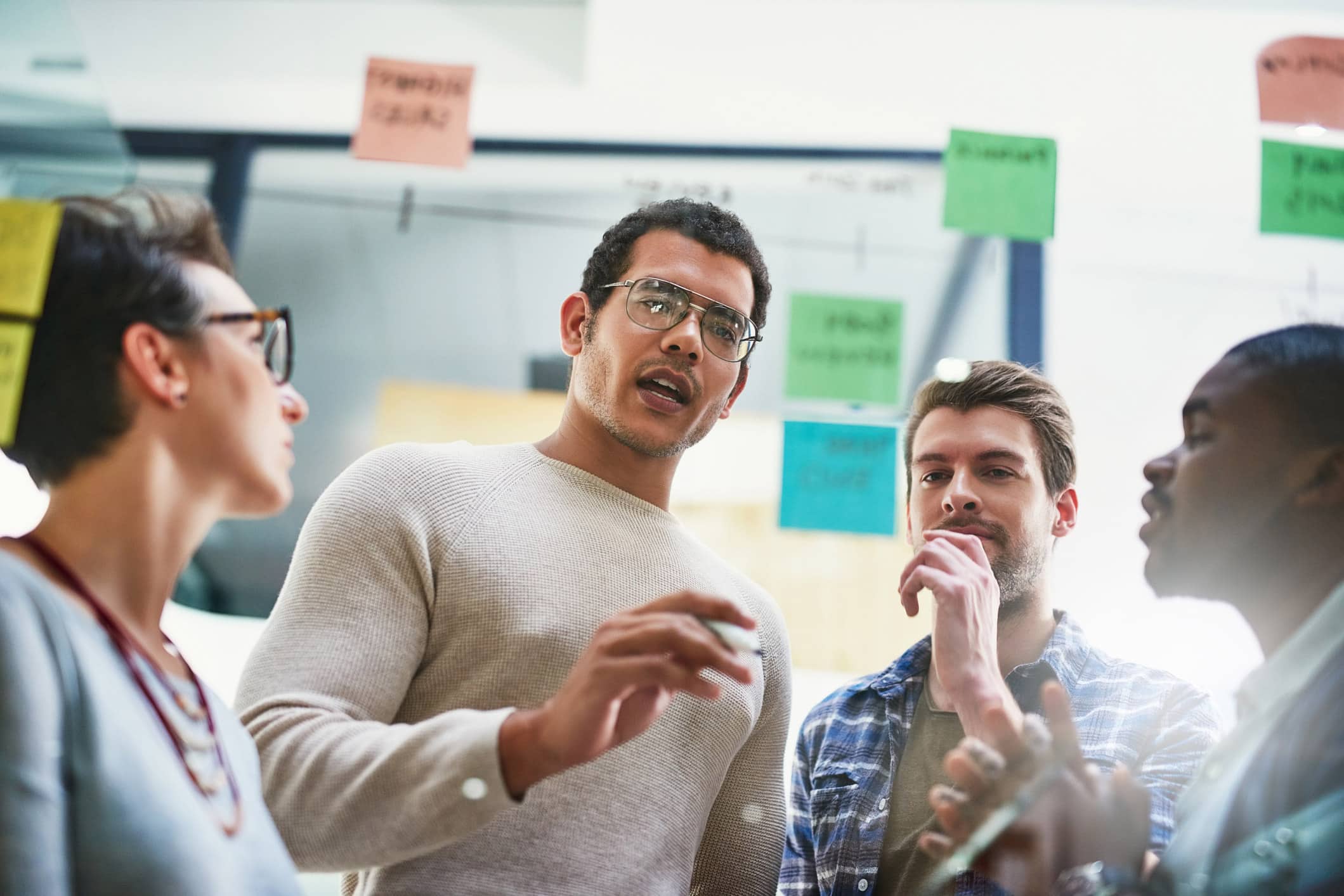 Four people discuss M&A Integration, standing by a window with colorful sticky notes behind them.