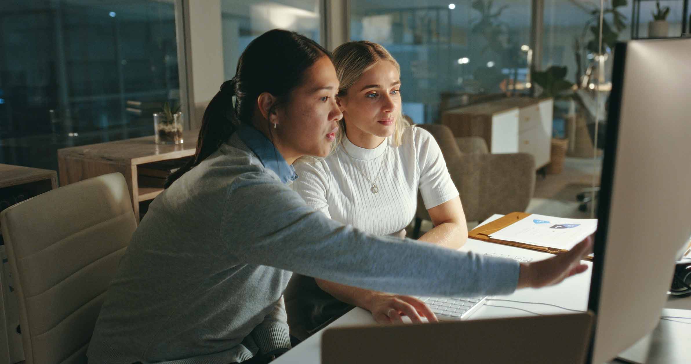 Two women working together at a computer in an office, one pointing at the screen.