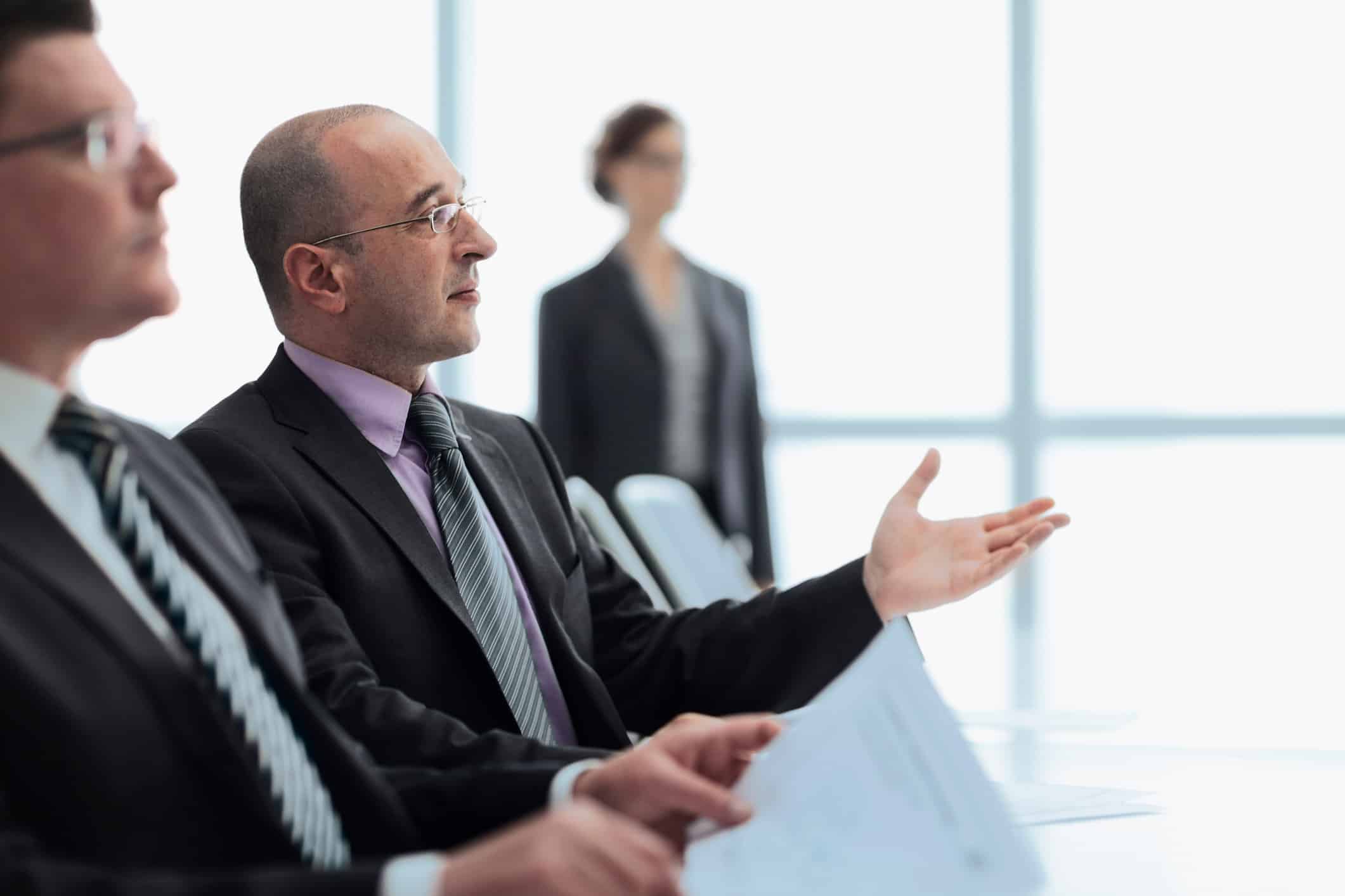 Man in suit speaks at meeting for forensic litigation consulting firm, papers in hand.