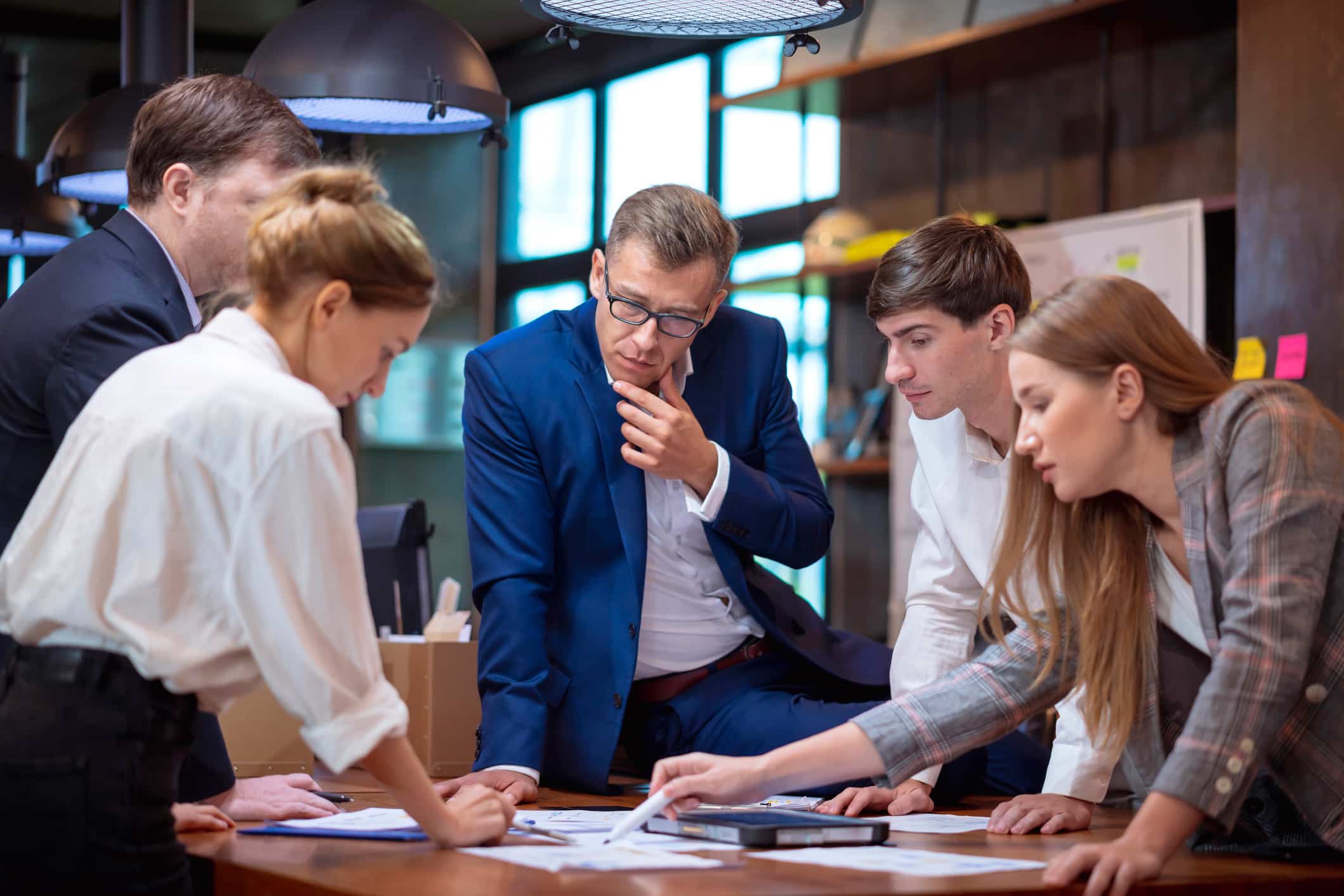 Five people in business attire discuss documents with Law Firm Growth Consultants in an office.