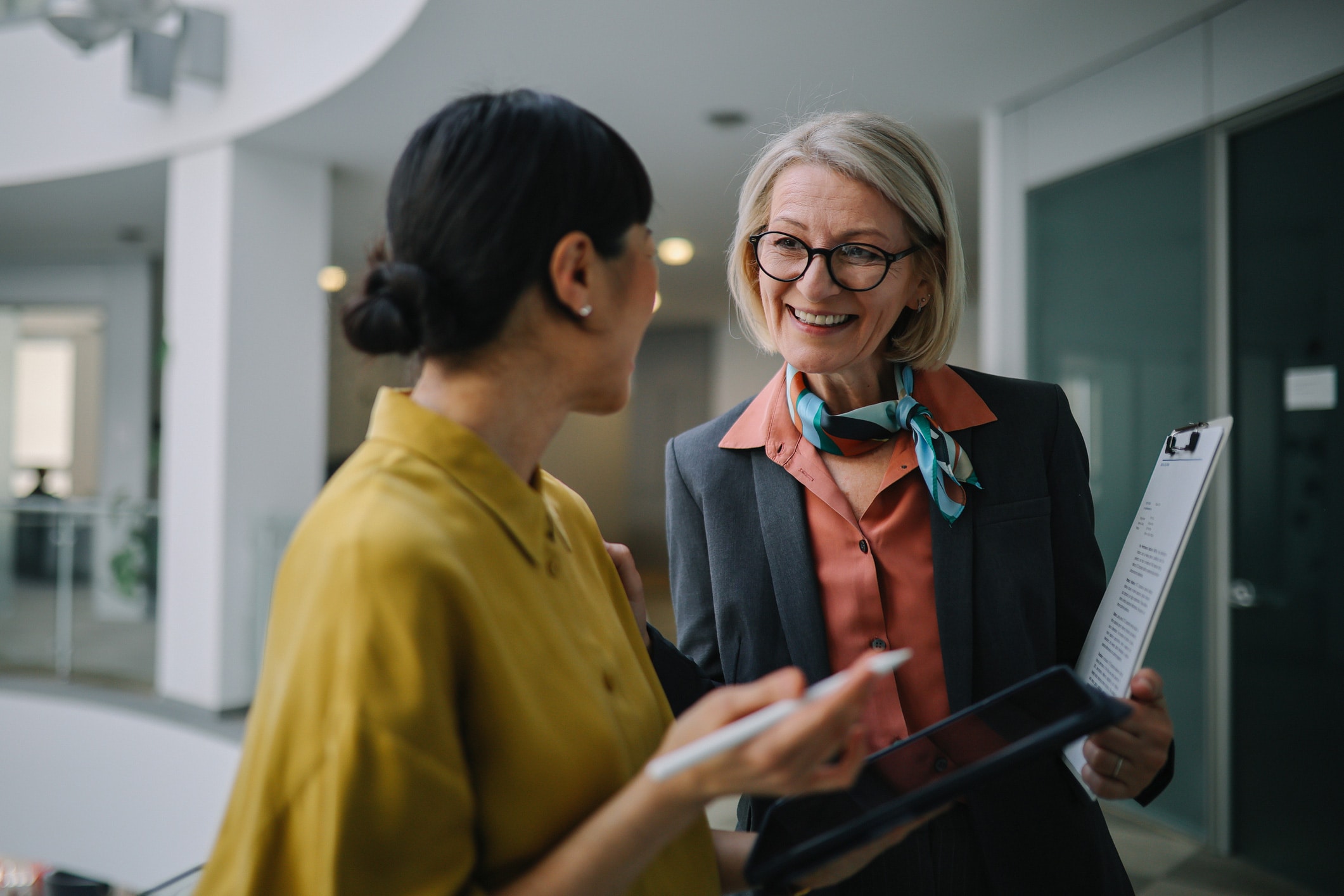Two women in business attire discuss Competitive Strategy Mapping, with a clipboard and tablet.