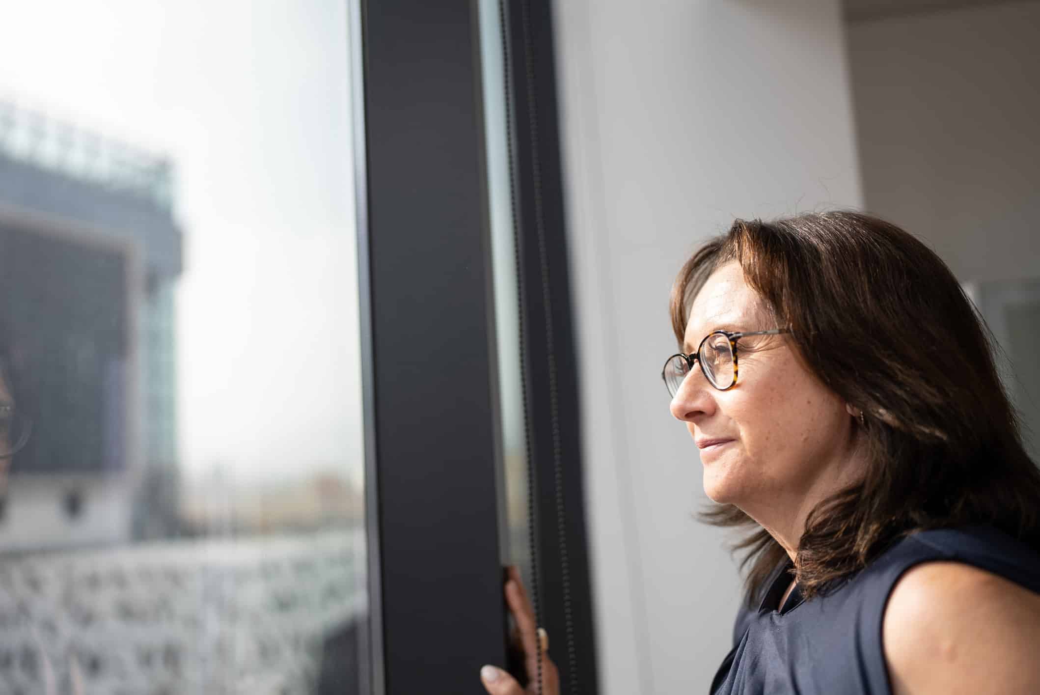 Woman with glasses looks thoughtfully out a window, reflecting on Witness Preparation Consulting.