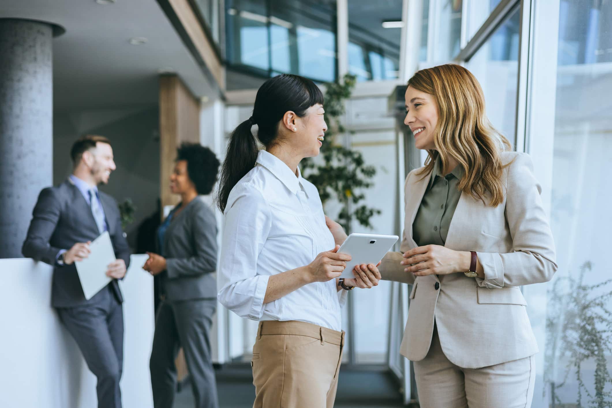 Two women discuss a Competitive Strategy Model in an office, colleagues chat in the background.