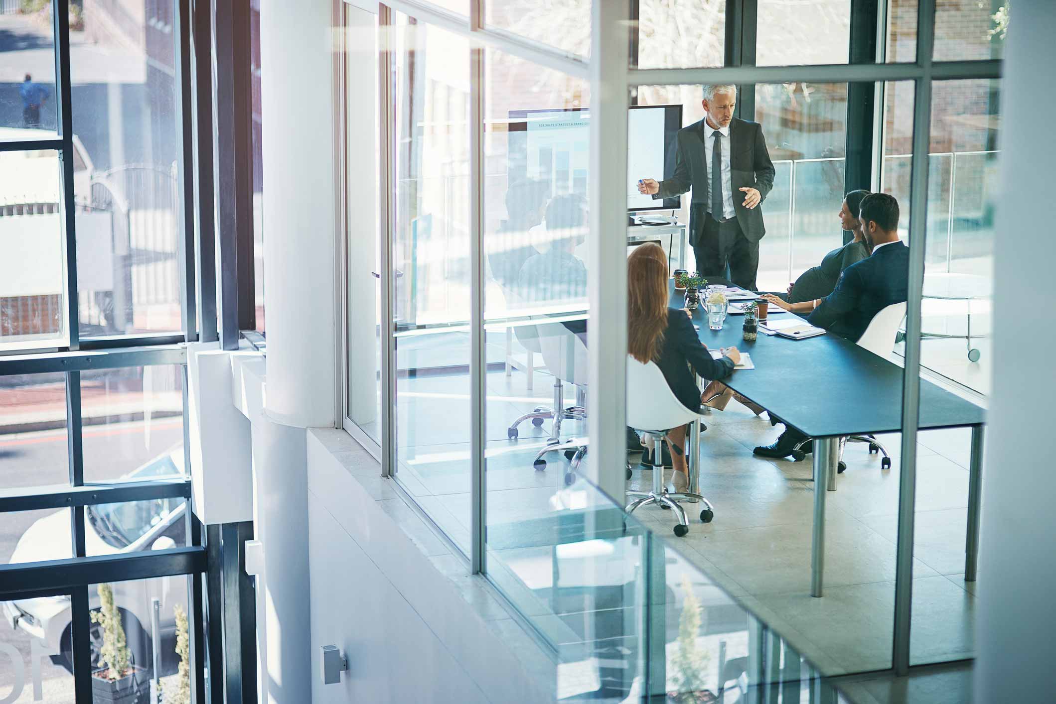 Business meeting in a modern glass office with three seated people and one person presenting.
