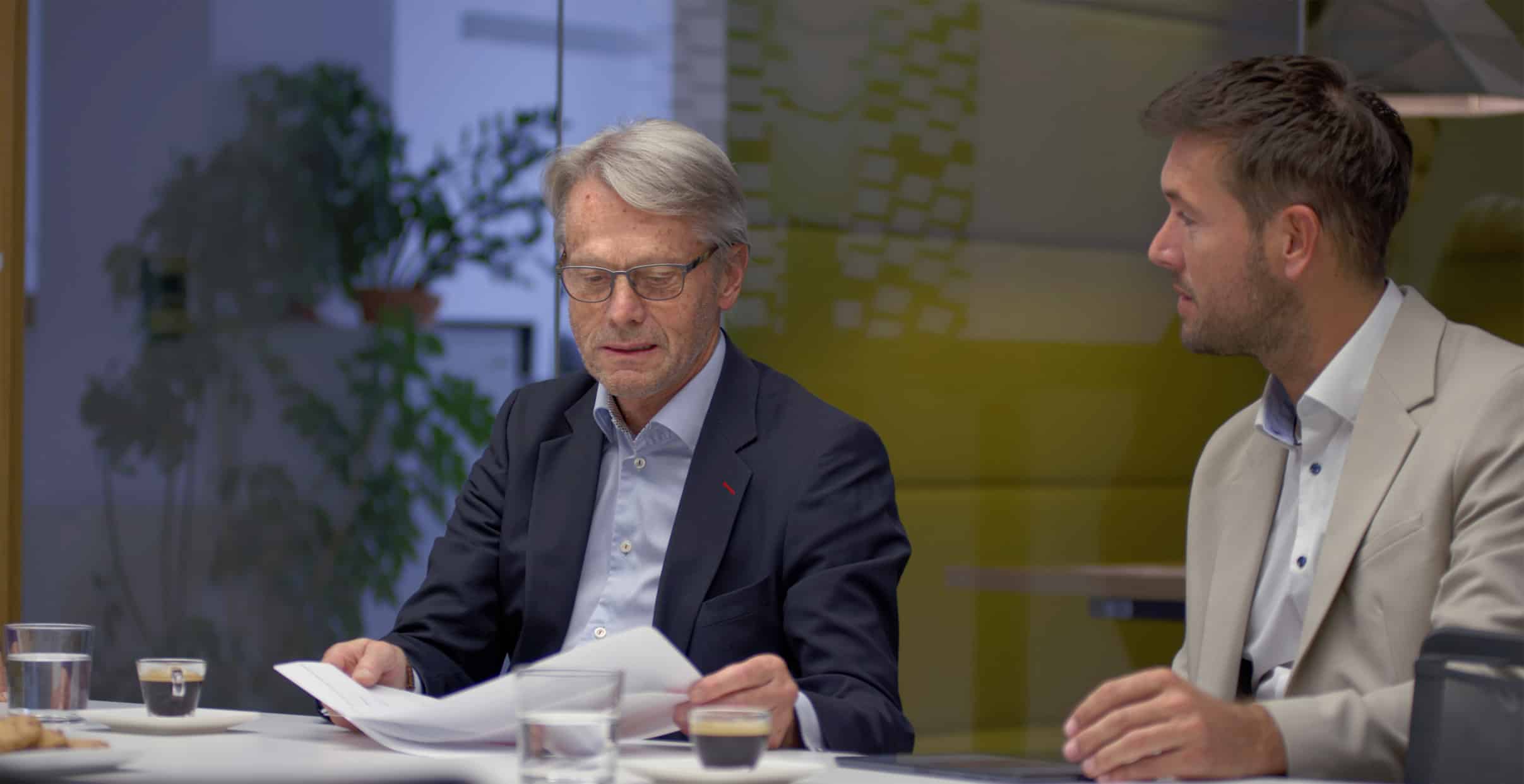 Two men in suits review documents and discuss Corporate Tax Advisory Services over coffee.
