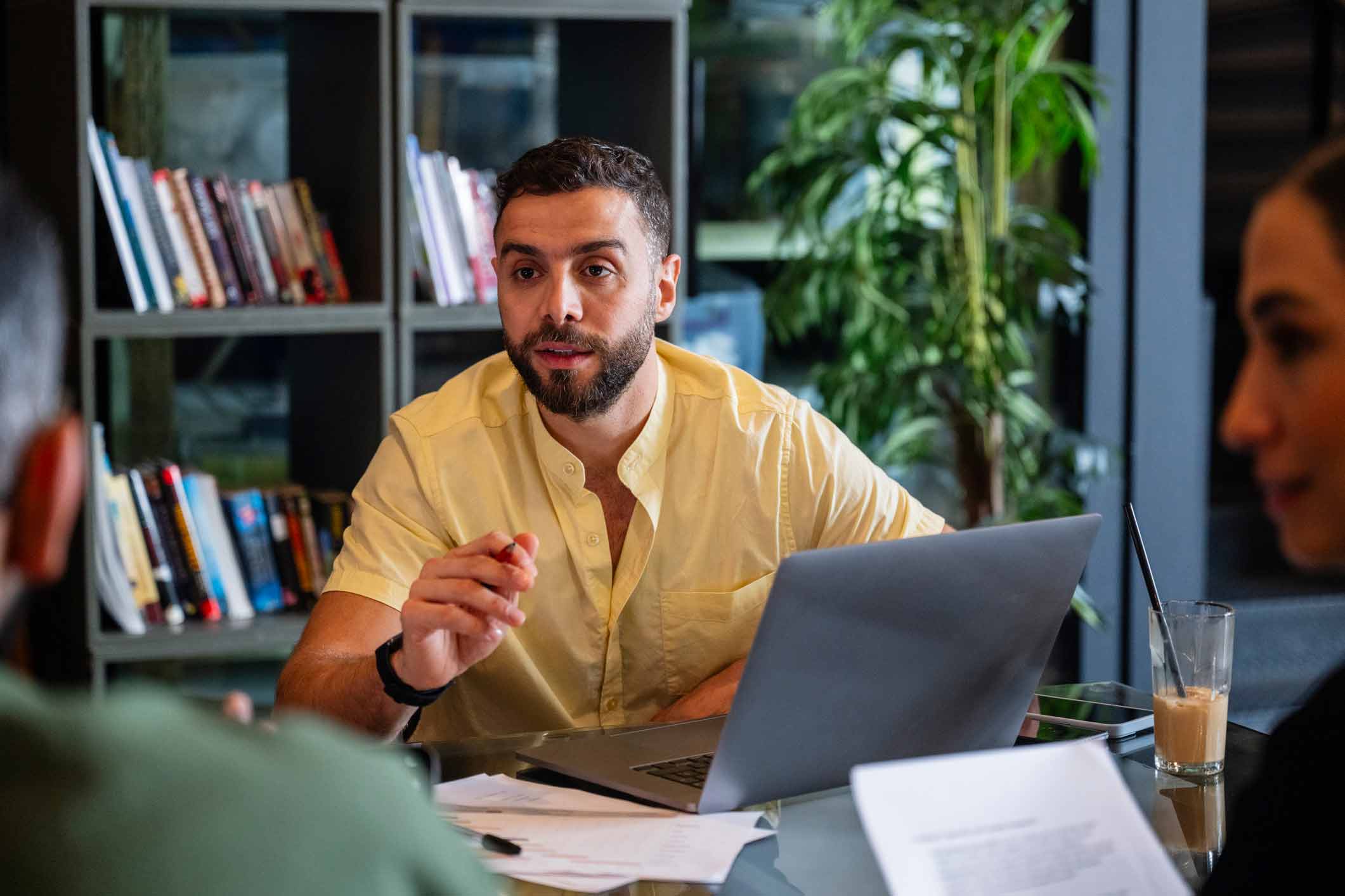 Man in yellow shirt discussing eDiscovery optimization at table with laptop, papers, & bookshelves.