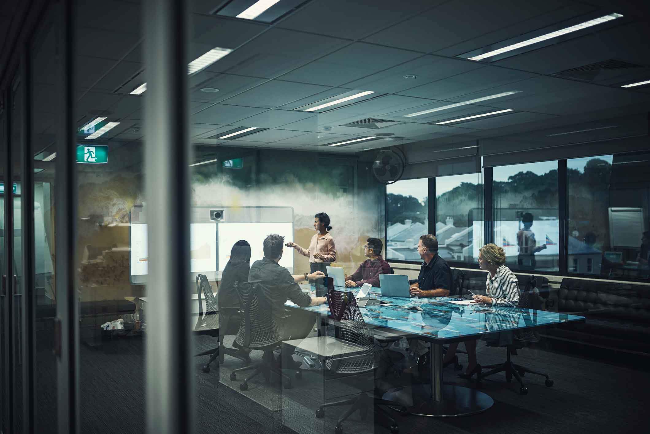 A woman presents on information security policy in a modern glass-walled conference room.